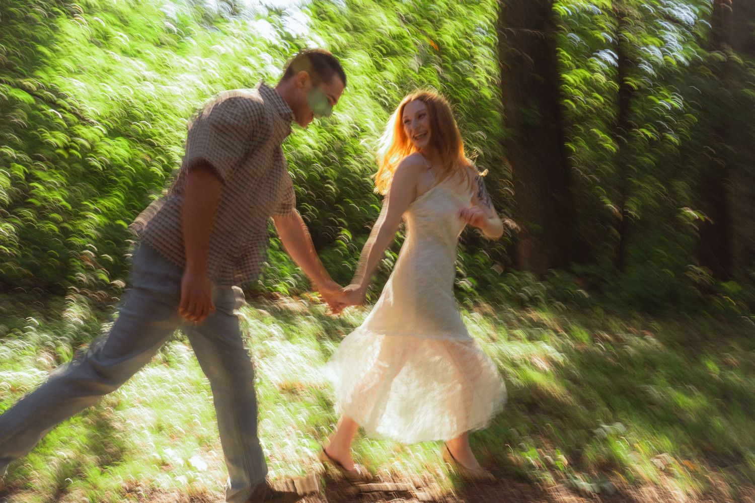 A couple holds hands and runs through a sunlit forest, the woman wearing a white dress in a dreamy blurred motion photo.