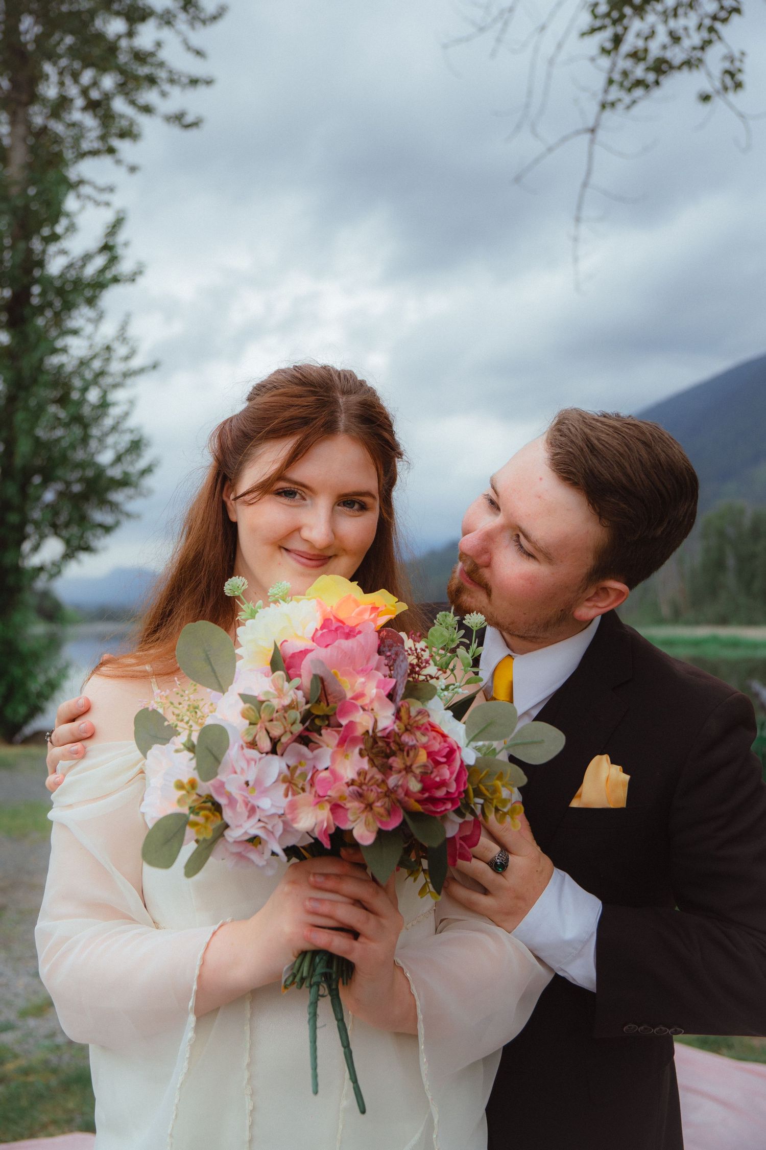 Bride holding colorful bouquet with groom embracing her outdoors near mountain lake on cloudy day.