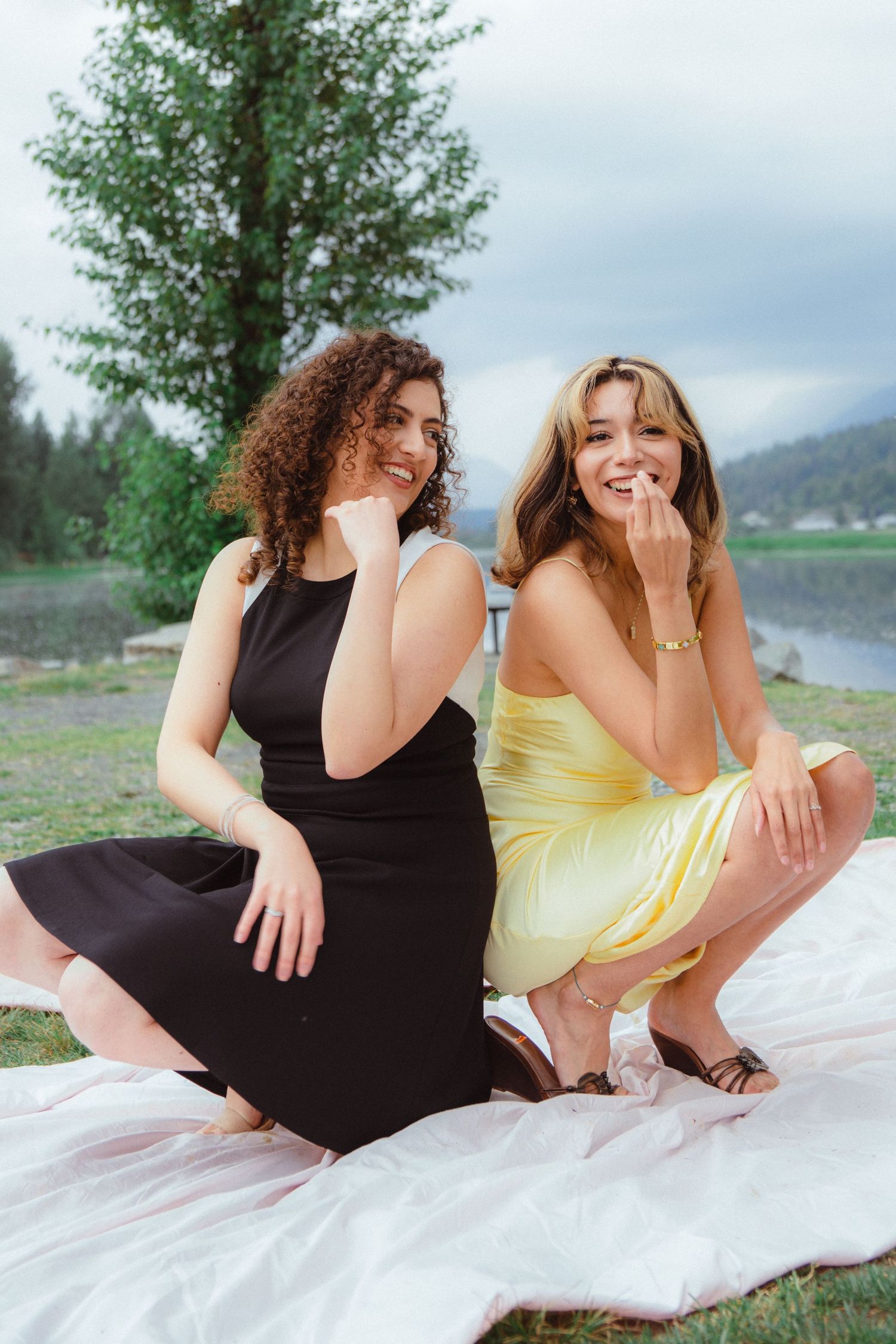 Two happy women in black and yellow dresses laugh together while sitting on a white blanket outdoors.
