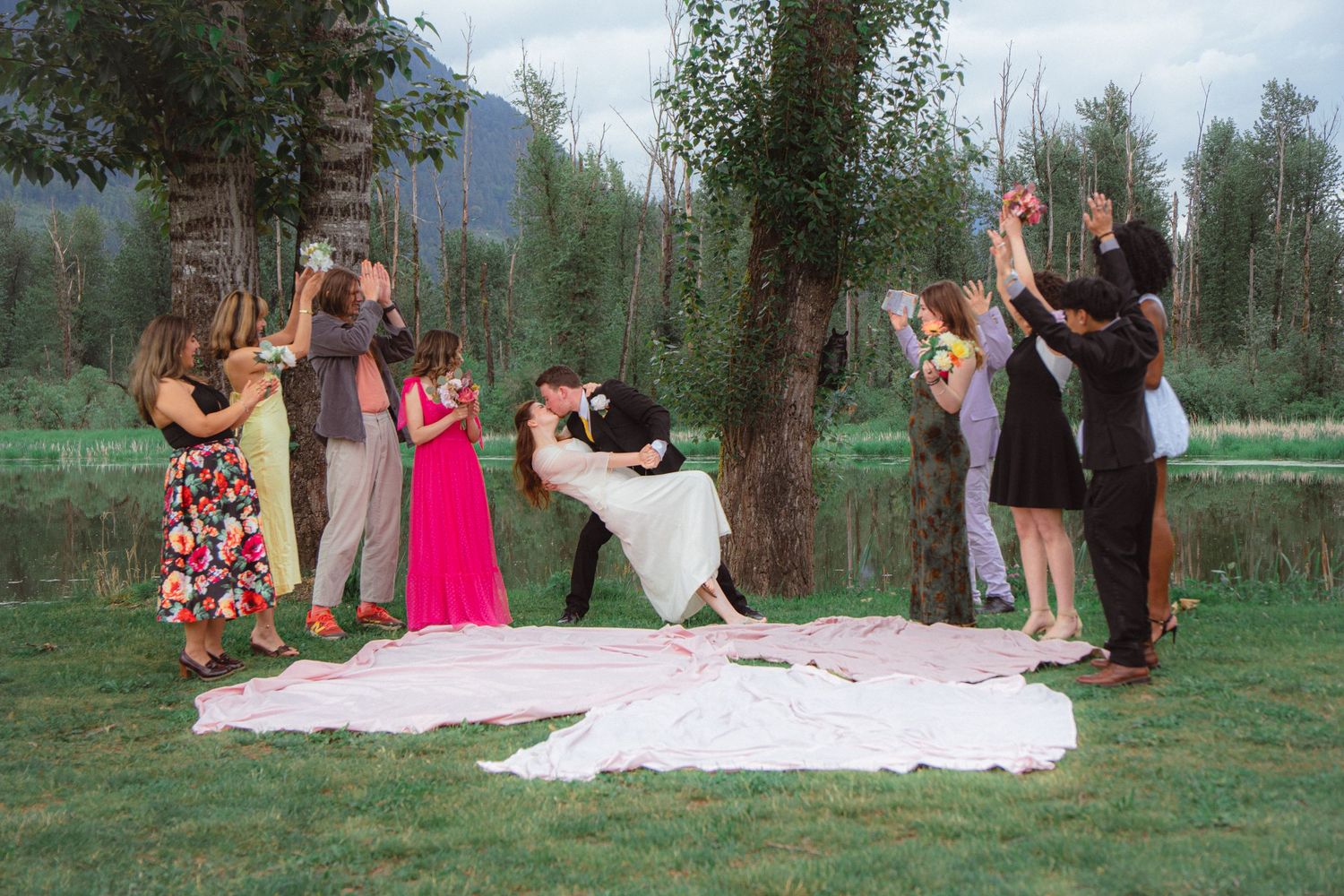 Groom dips bride romantically as cheering wedding guests celebrate beside a scenic lakeside setting.
