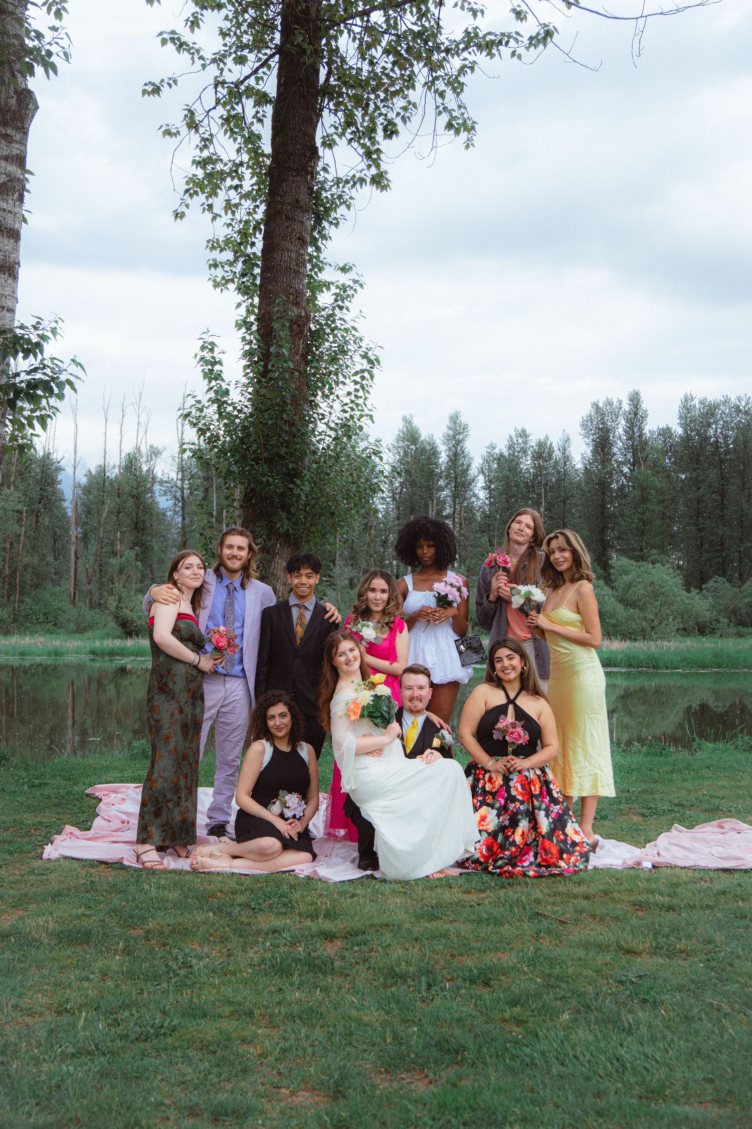 Bride and groom surrounded by wedding party in colorful formal attire near a lakeside tree.