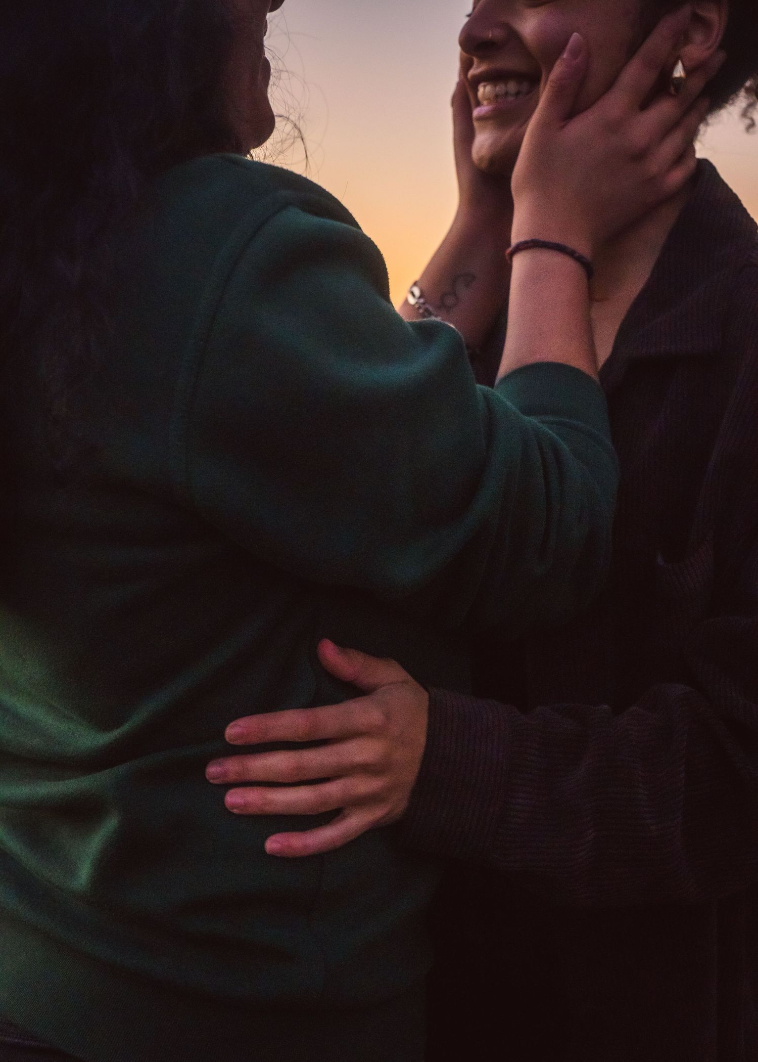 Two women share an affectionate embrace at sunset, one smiling brightly as her partner holds her face tenderly.