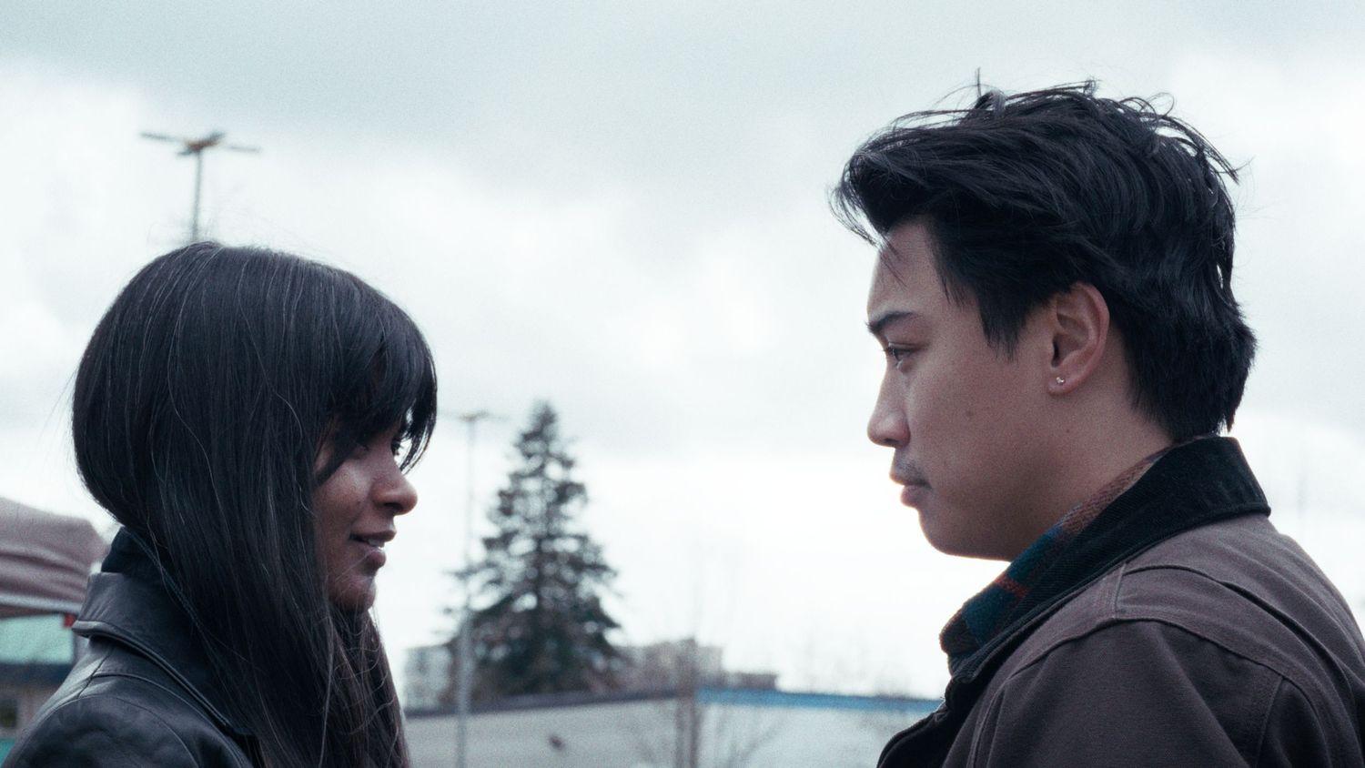 Young Asian man and woman face each other outdoors under overcast sky.