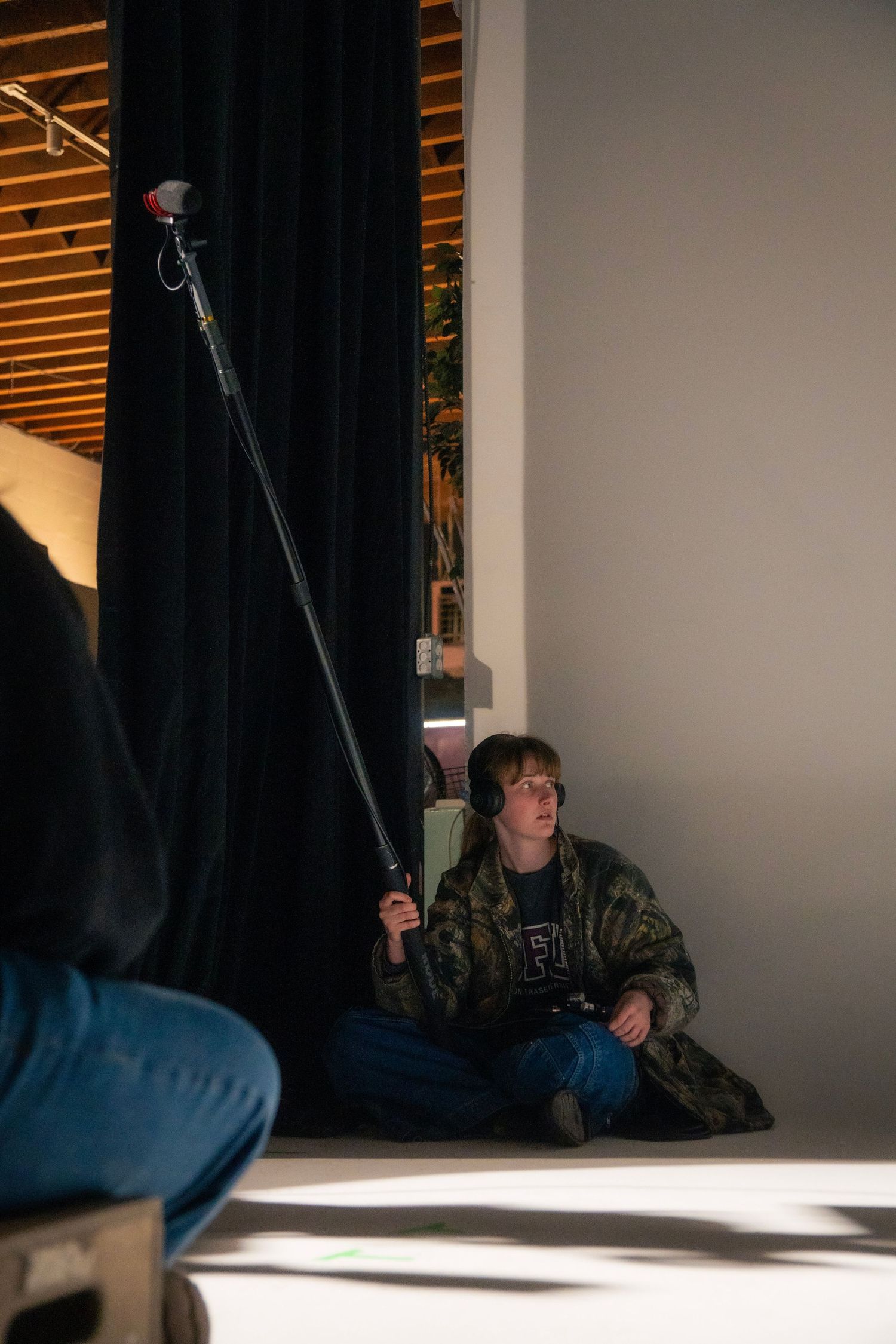 Person sitting on floor holding boom microphone pole with headphones in a film production setting.