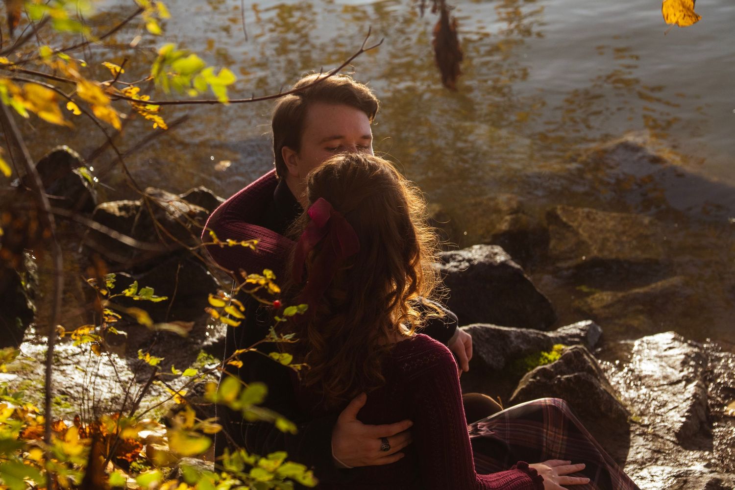 Couple sharing intimate moment sitting on rocks by a sunlit autumn lake surrounded by fall foliage.