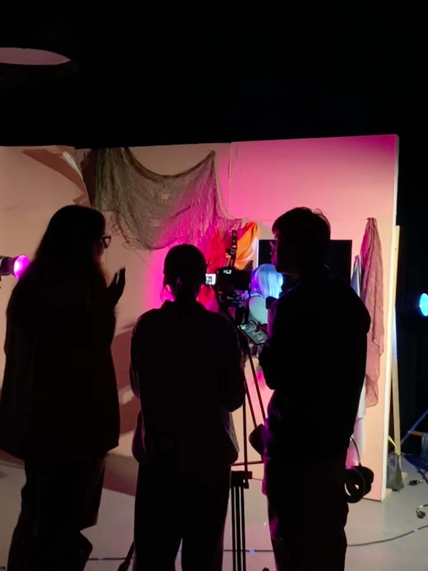 Three silhouetted figures operating a camera on a film set with pink and purple lighting.