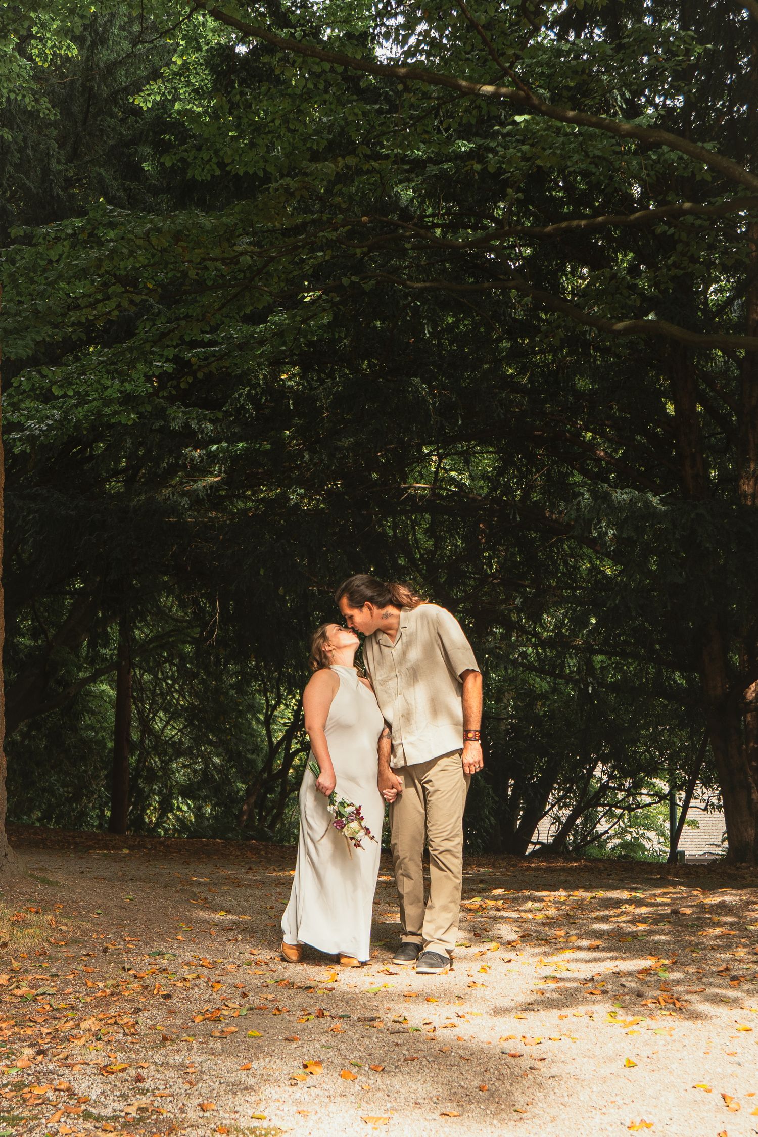 Bride and groom sharing a tender kiss on a sunlit forest path surrounded by lush green trees.