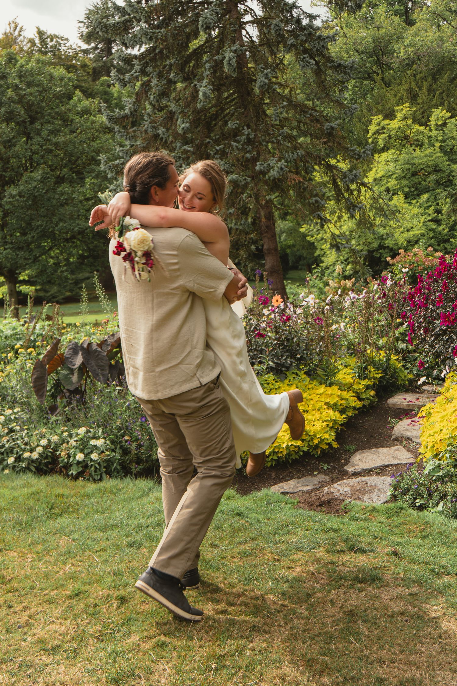 Happy bride and groom embracing in a garden, groom lifting bride surrounded by vibrant flowers and greenery.