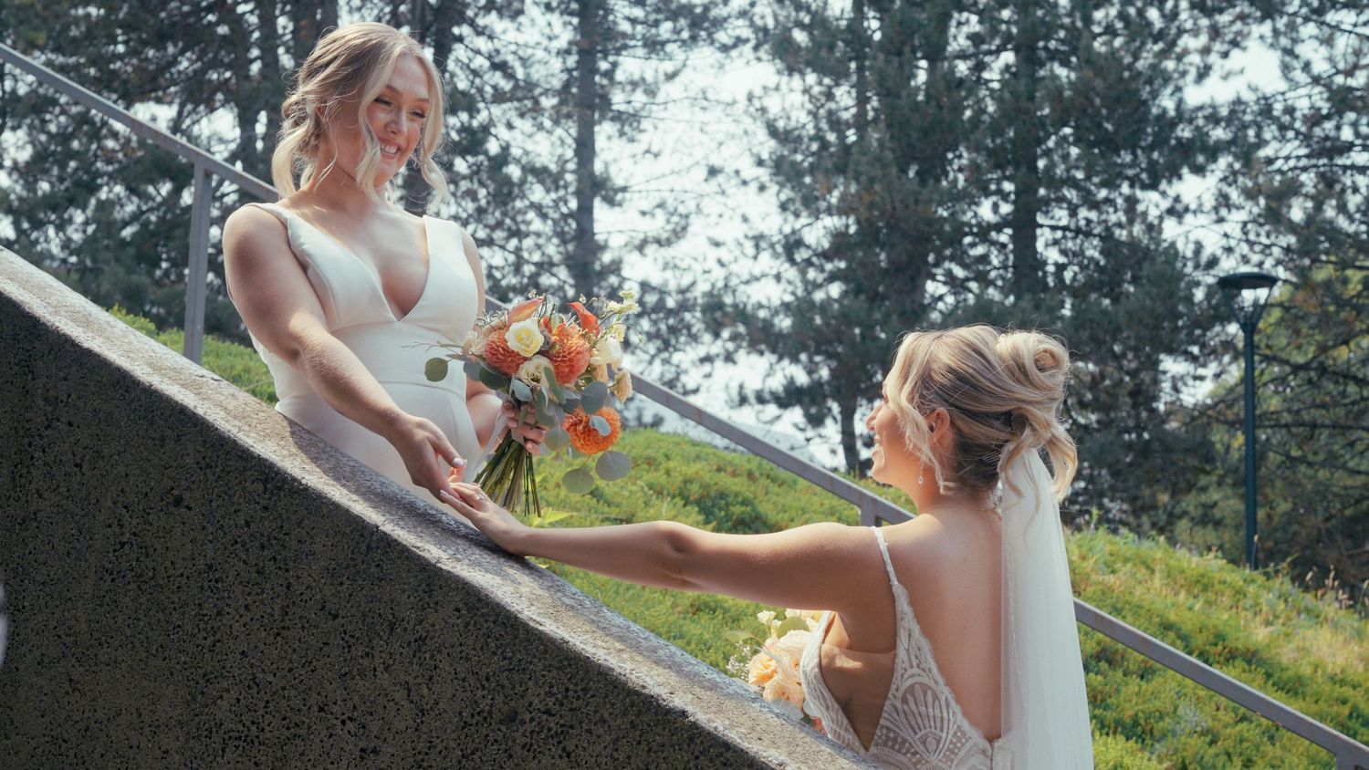 Two brides exchange a bridal bouquet on outdoor stone steps surrounded by lush greenery.