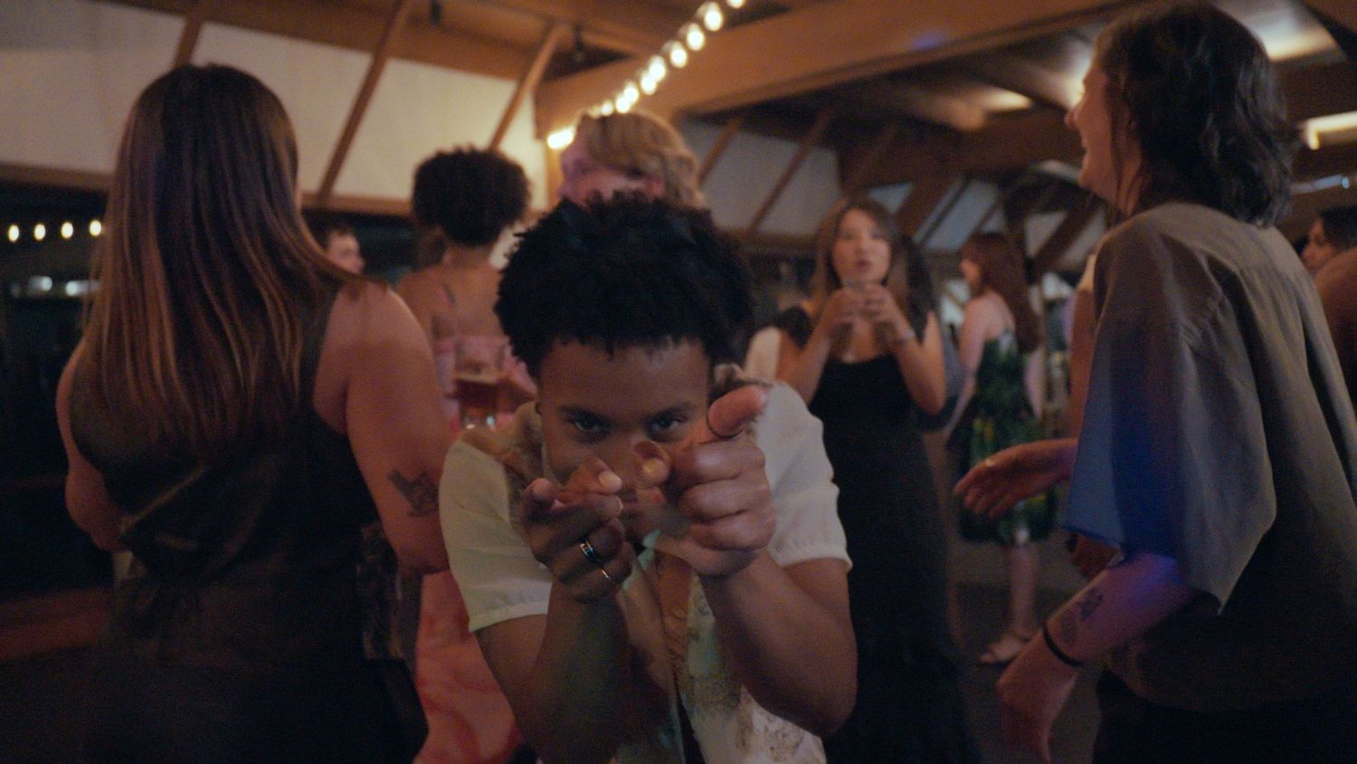 Young man dancing at a lively indoor party, pointing at camera, with guests socializing in warm ambient lighting.