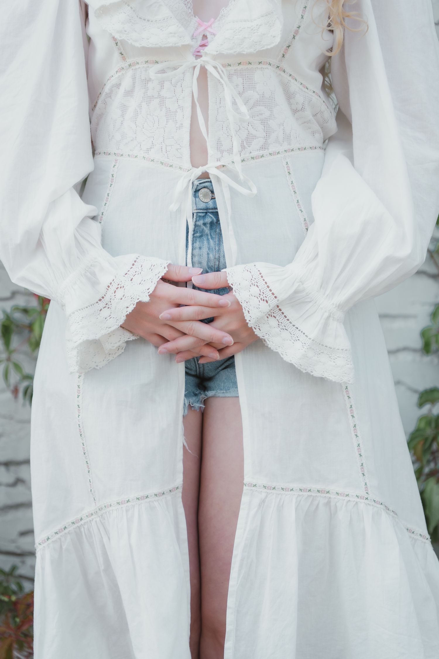 Woman wearing open white lace boho duster coat over denim shorts, hands clasped at waist.