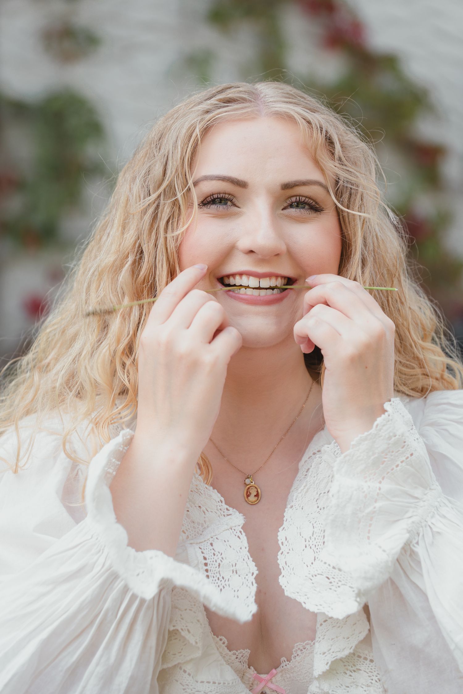 Smiling blonde woman in white lace blouse with curly hair holding face, wearing gold pendant necklace.