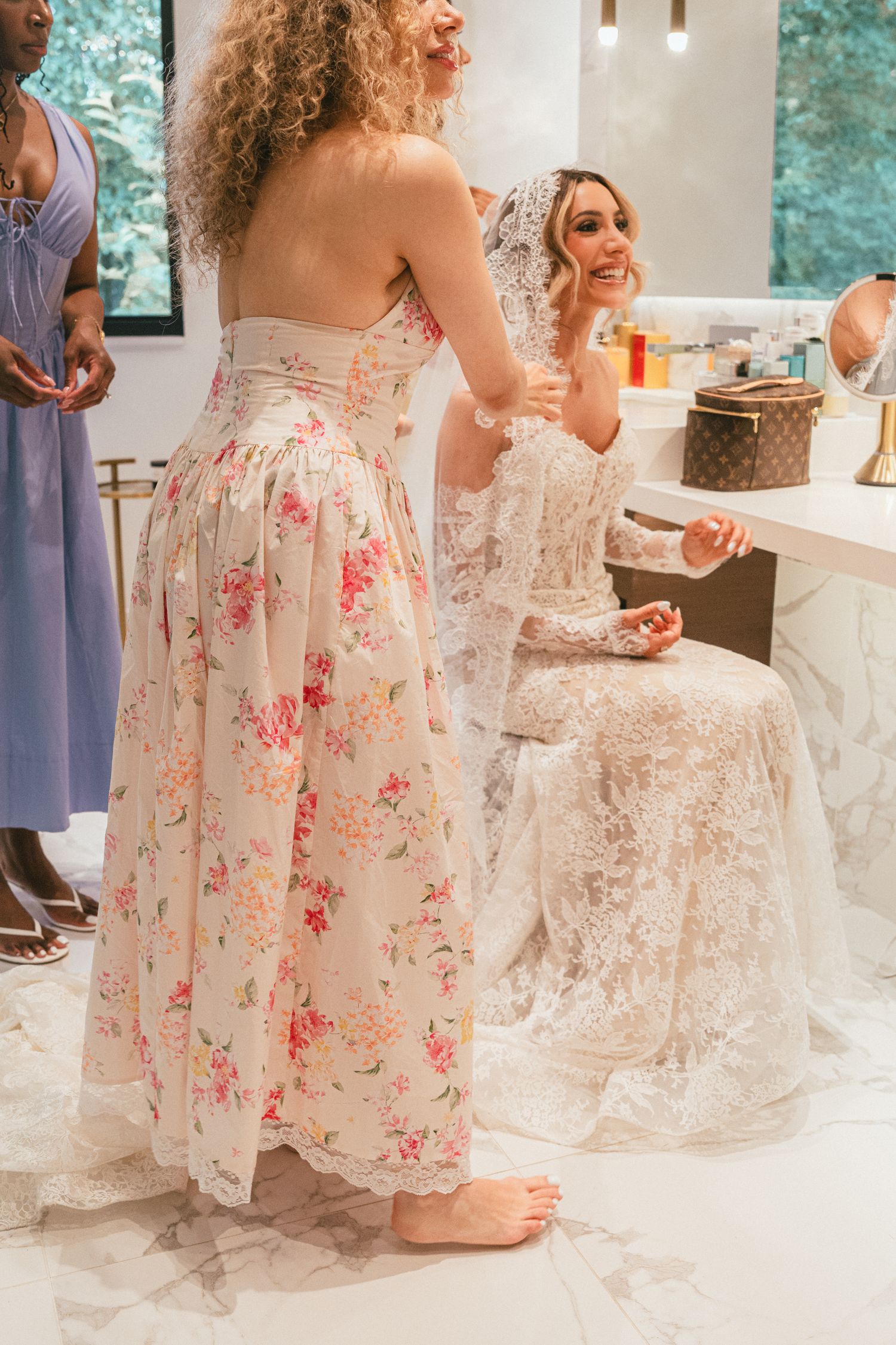 Bridesmaid in floral gown assisting the bride with her lace wedding dress at the vanity mirror.
