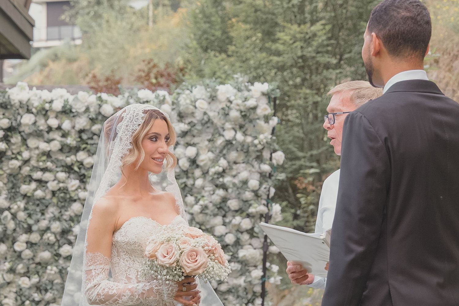Bride in lace gown and veil holds roses bouquet during outdoor wedding ceremony with floral backdrop.