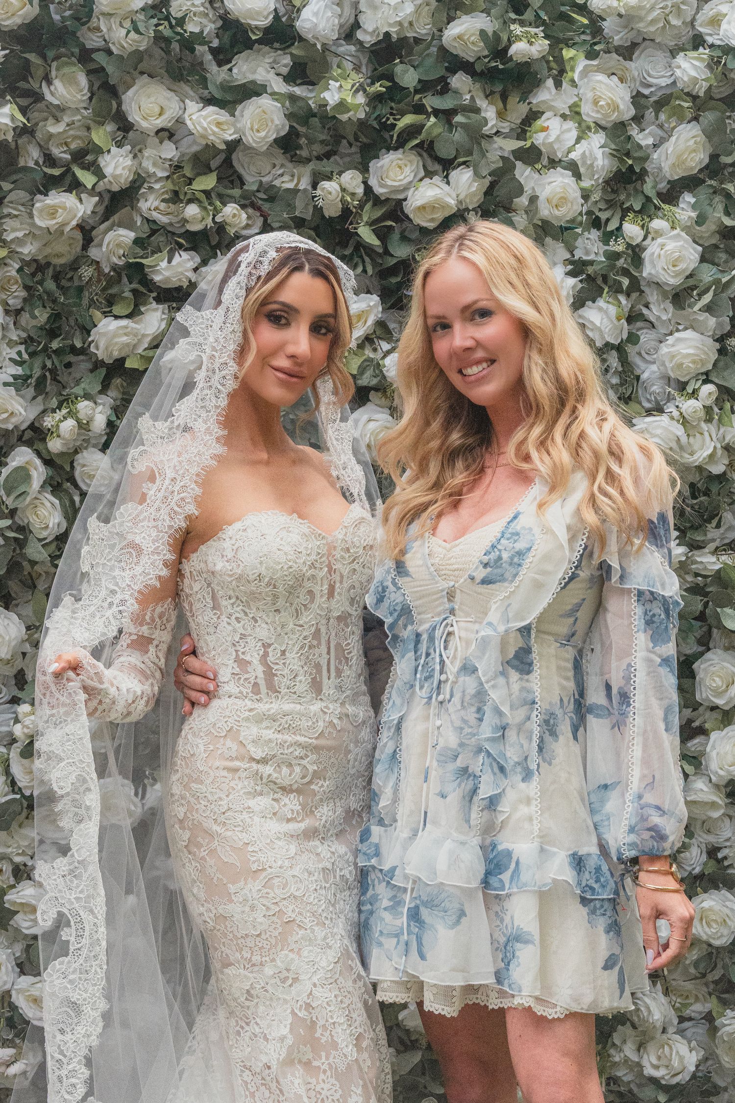 Bride in lace wedding gown and veil poses with blonde woman in floral dress before white rose wall backdrop.