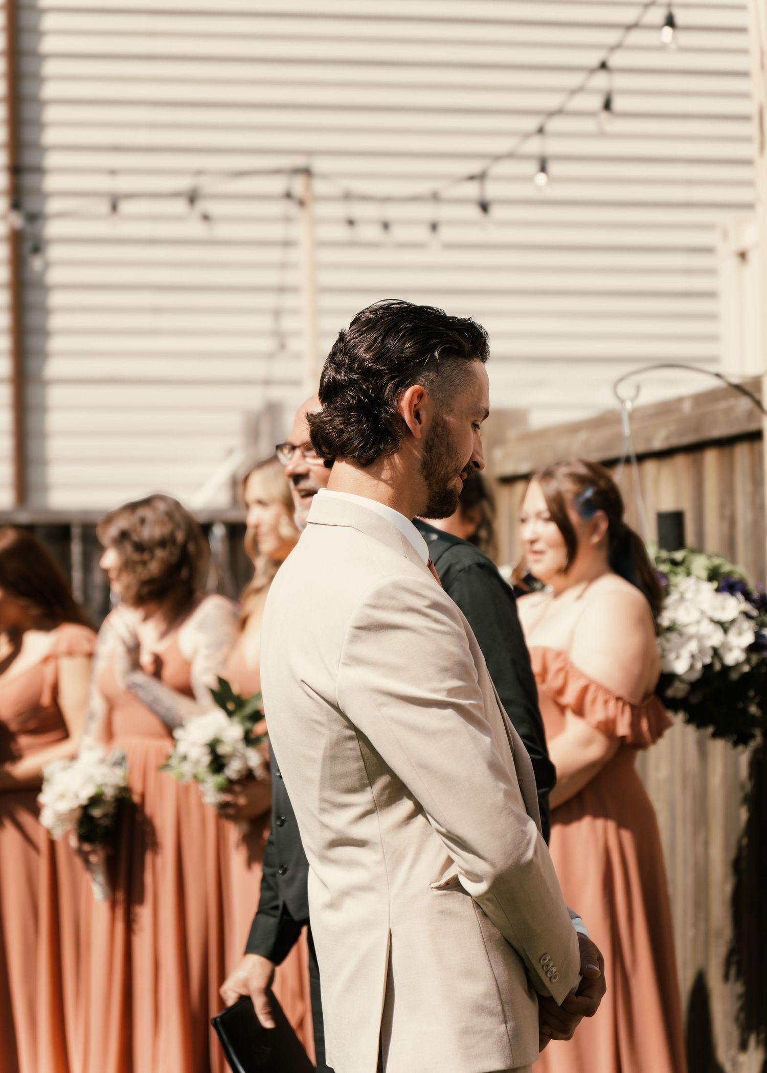 Groom in beige suit stands at outdoor wedding altar with bridesmaids in terracotta dresses holding bouquets behind him.