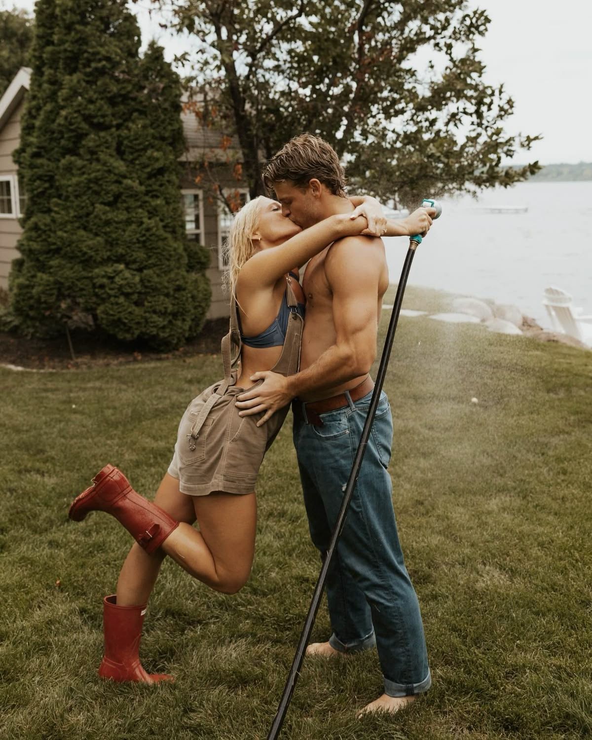 Couple sharing a romantic kiss outdoors by a lakeside yard, woman wearing red rain boots and man holding a long pole.
