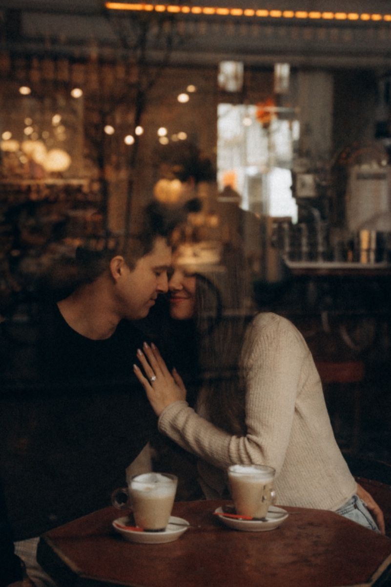 Romantic couple sharing an intimate moment over lattes in a warmly lit cafe, viewed through glass.