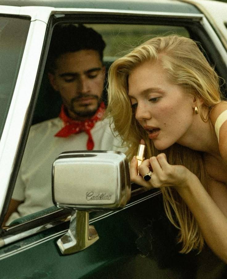 A blonde woman applies lipstick using a Cadillac side mirror while a man in a red bandana watches from inside the car.