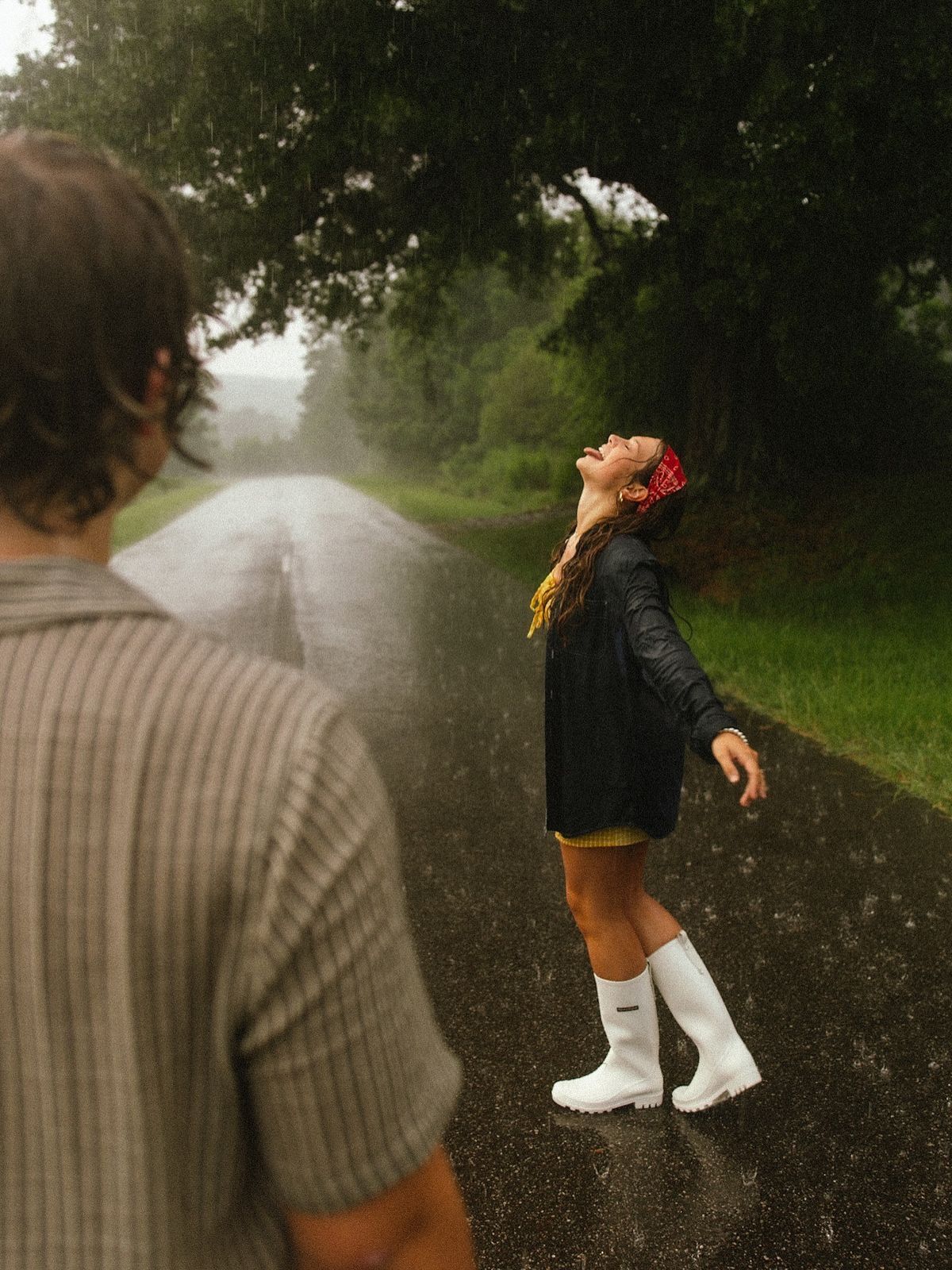 Woman in white boots and black jacket joyfully tilts head back in the rain on a misty tree-lined road.