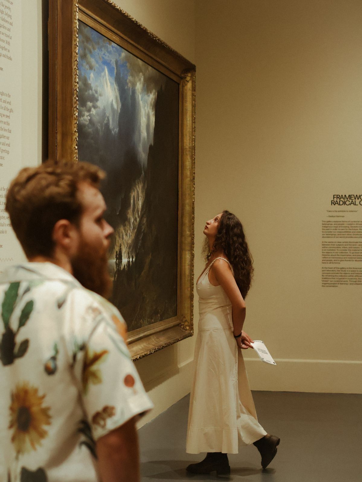 Woman in white dress gazes up at a large landscape painting in a museum gallery, man in floral shirt in foreground.