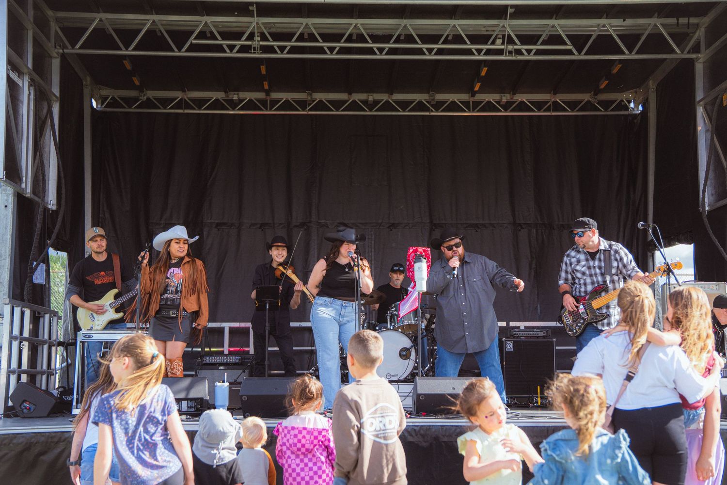 Country band performs on outdoor stage for crowd of adults and children at daytime music festival.