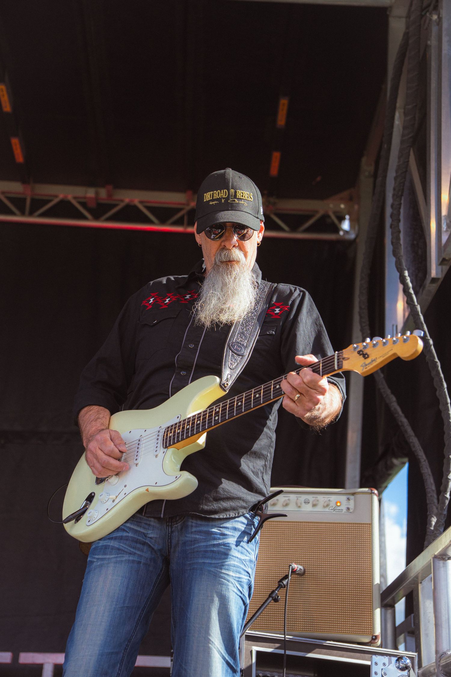 Bearded guitarist in black shirt and cap playing white Stratocaster on outdoor stage.