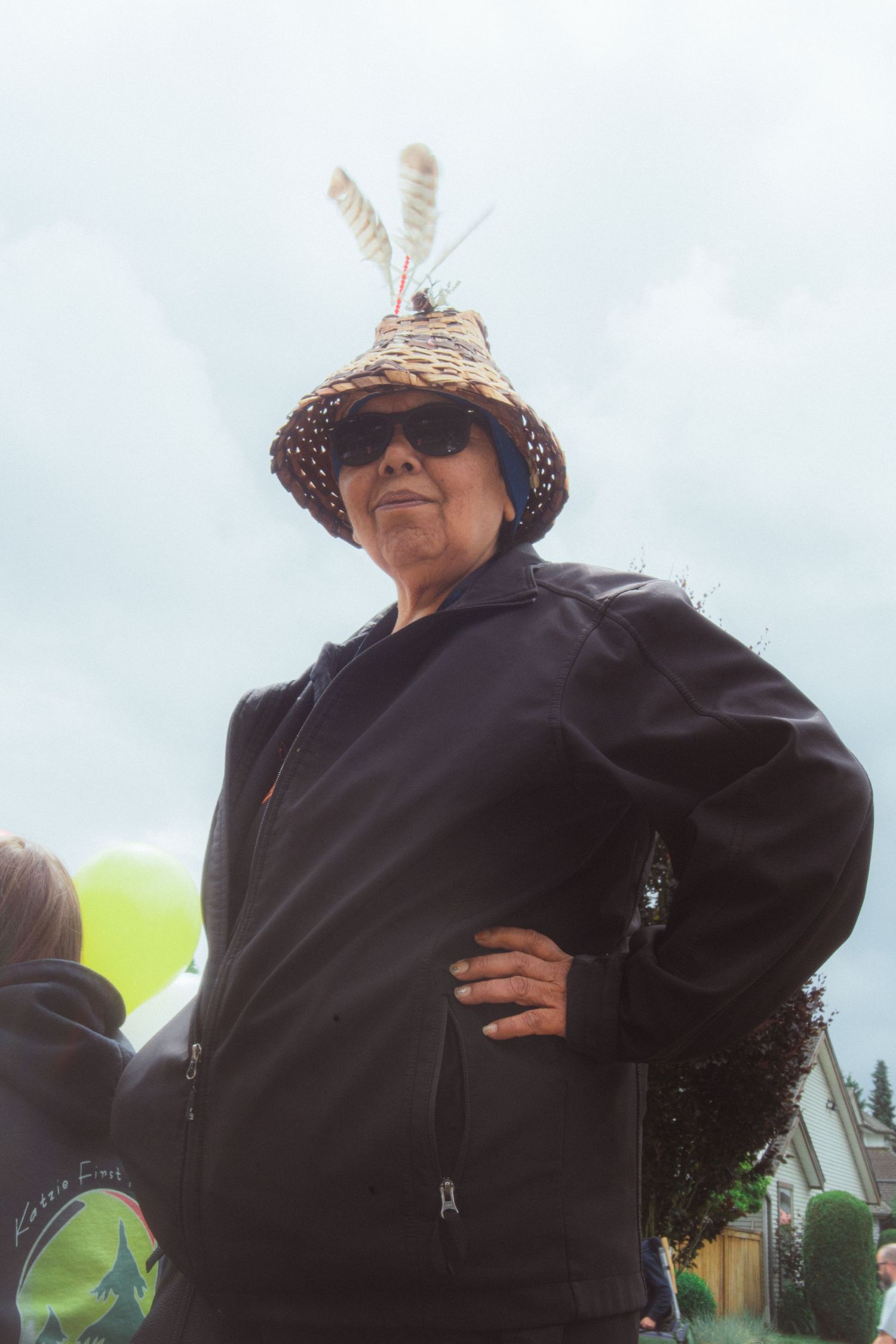 Indigenous woman wearing a traditional woven hat with feathers stands confidently at an outdoor event.