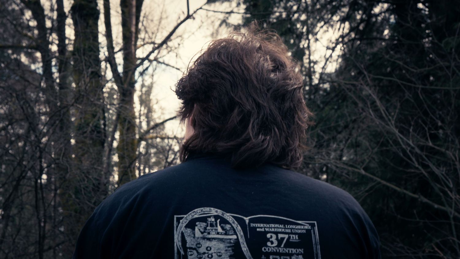 Back view of a person with long hair wearing a black graphic t-shirt standing in a wooded forest area.
