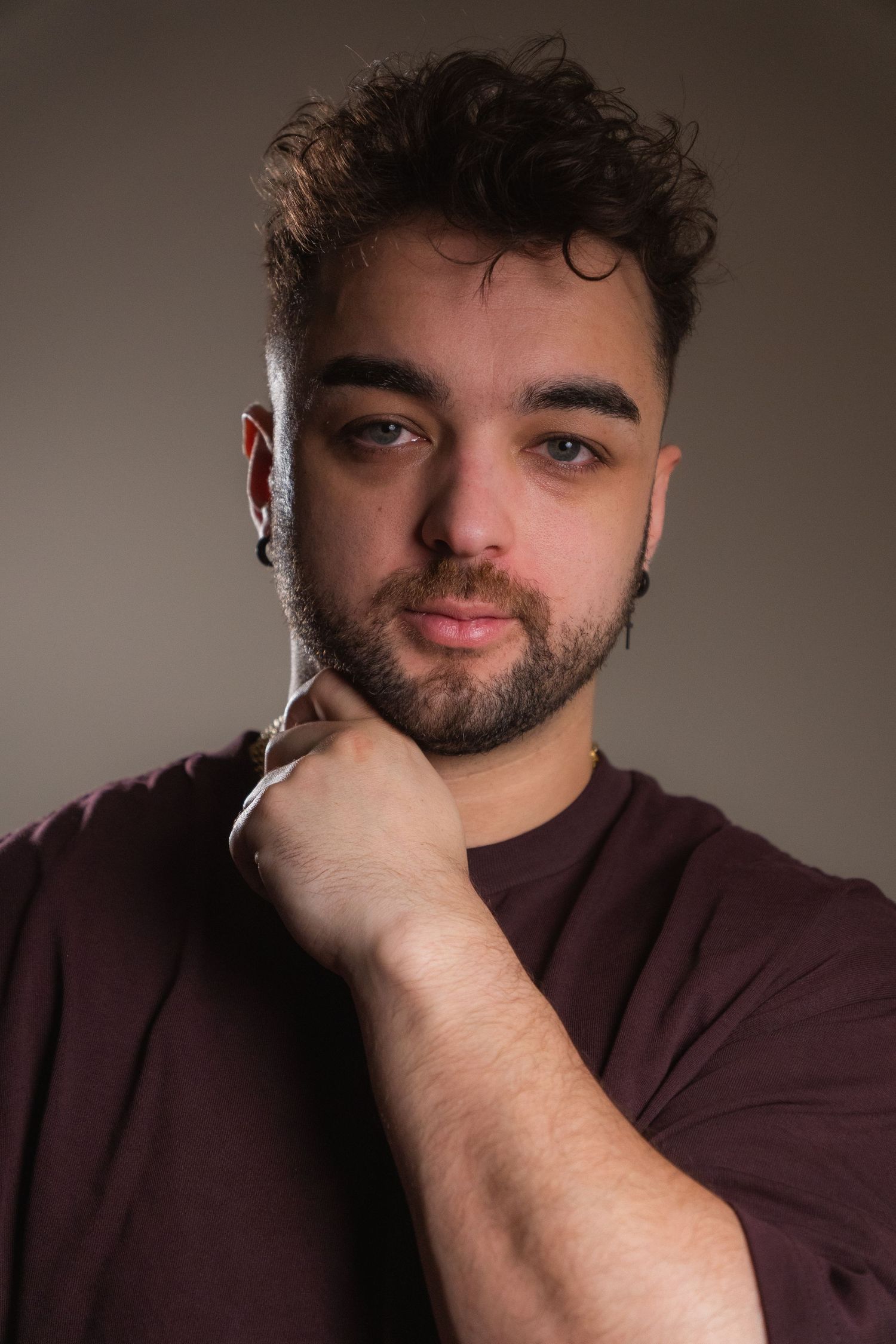 Young man with curly hair and beard wearing maroon shirt, posing with hand raised to chin against gray background.