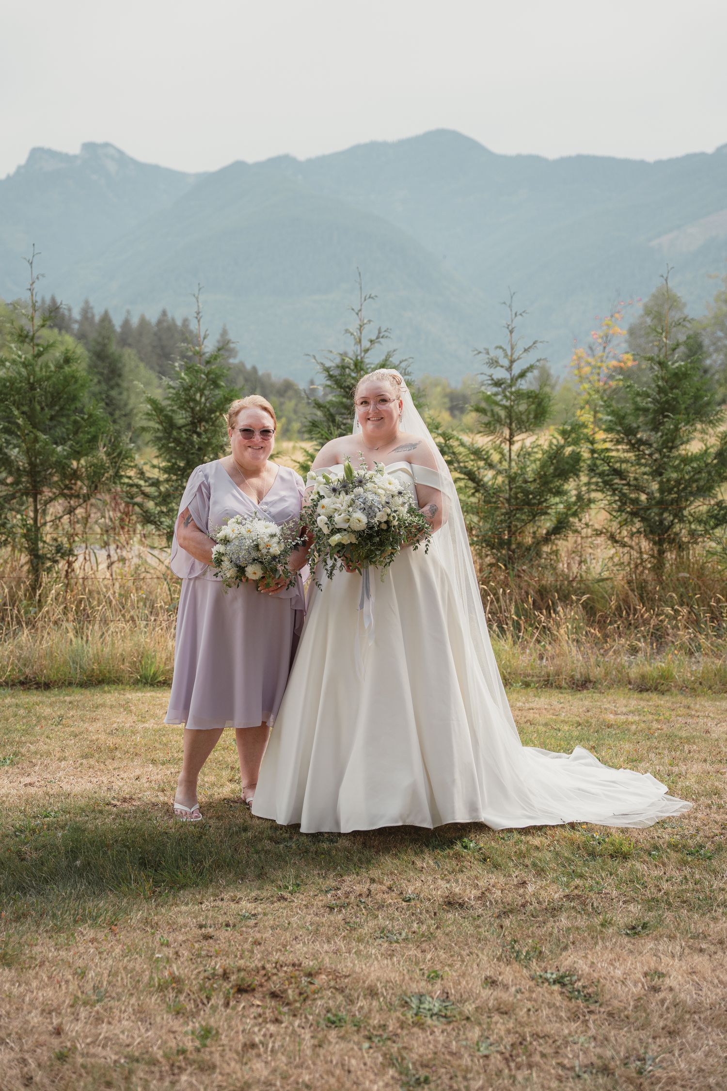 Bride in white gown and bridesmaid in lavender dress holding bouquets with mountain backdrop.