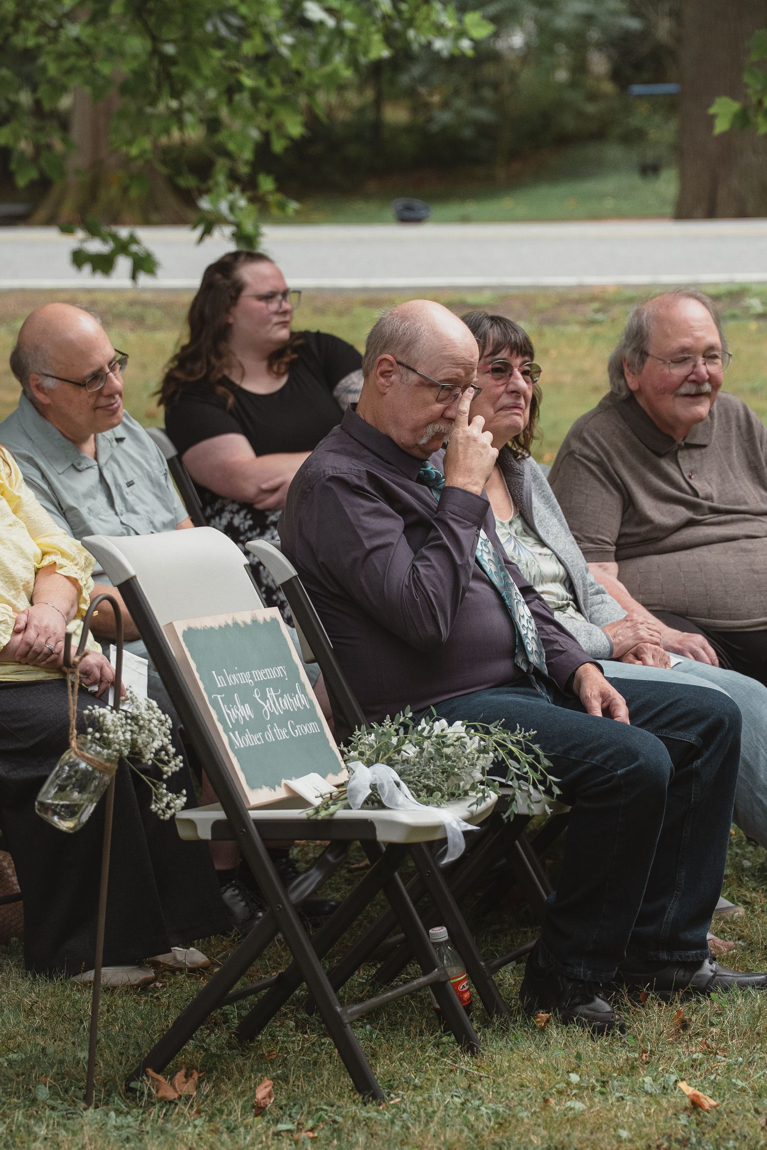 Wedding guests seated outdoors, emotional man wiping tears during ceremony with memorial chair.