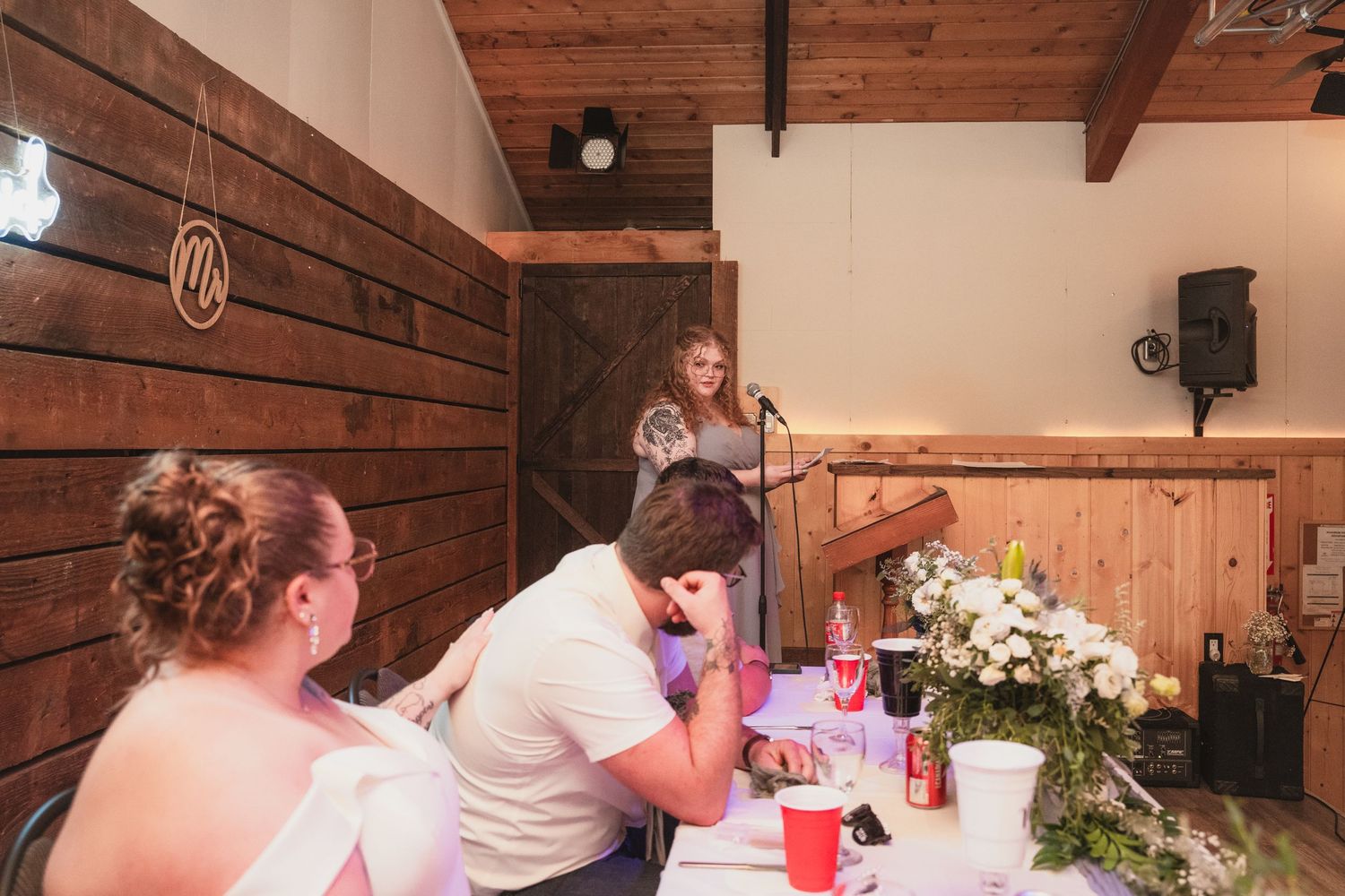 Wedding guest delivers a touching speech at the microphone during a rustic barn reception while guests listen.