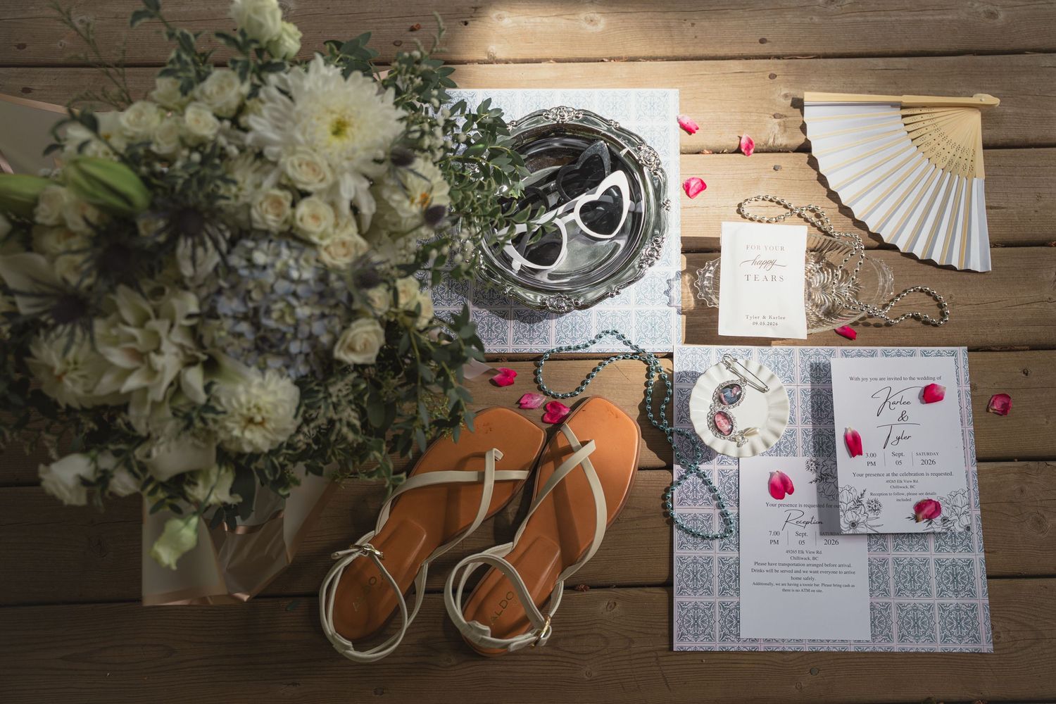 Flat lay of wedding accessories including sandals, bouquet, sunglasses, fan, jewelry, and invitation on wood.