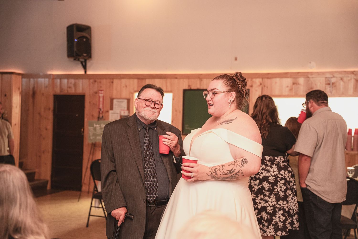 Bride in white gown chats with a guest holding red cups at a casual rustic wedding reception.