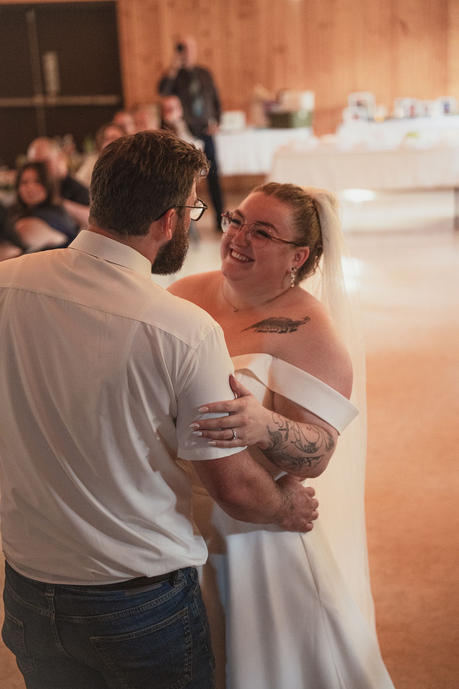 Smiling bride with tattoos shares first dance with groom in white shirt at wedding reception.