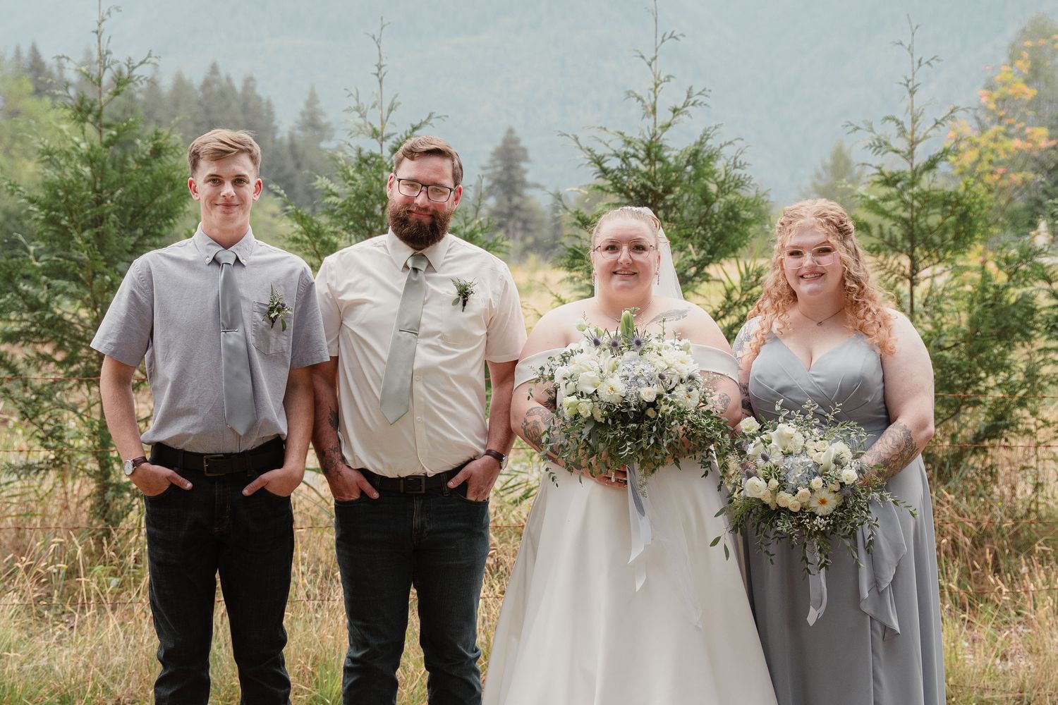 Wedding party posing outdoors with mountains, bride in white gown holding bouquet, bridesmaid in gray dress, two groomsmen.