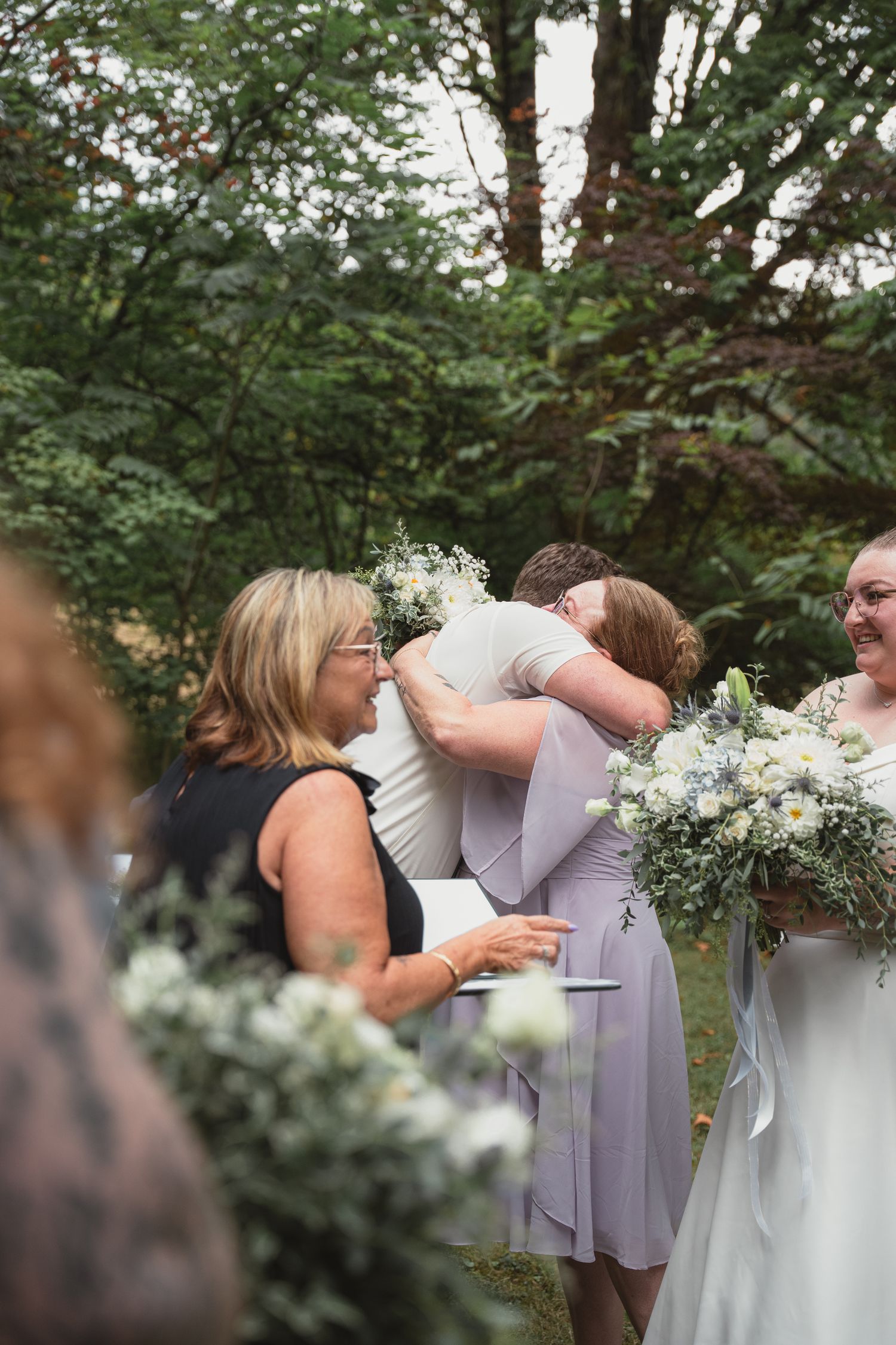 Bridesmaids in lavender dresses share a warm embrace while holding bouquets at an outdoor wedding ceremony.