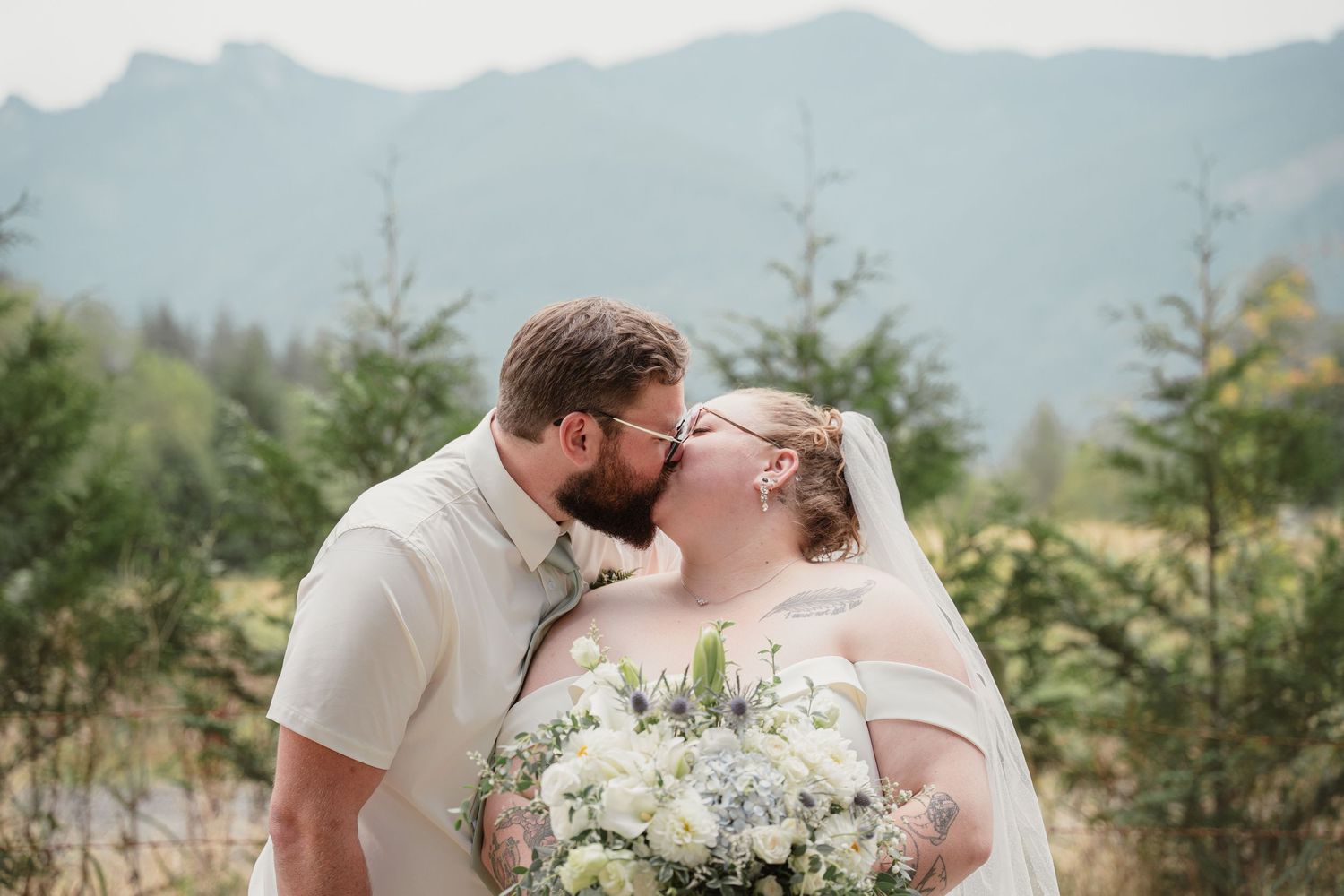 A bride and groom share a kiss outdoors with lush green trees and misty mountains in the background on their wedding day.