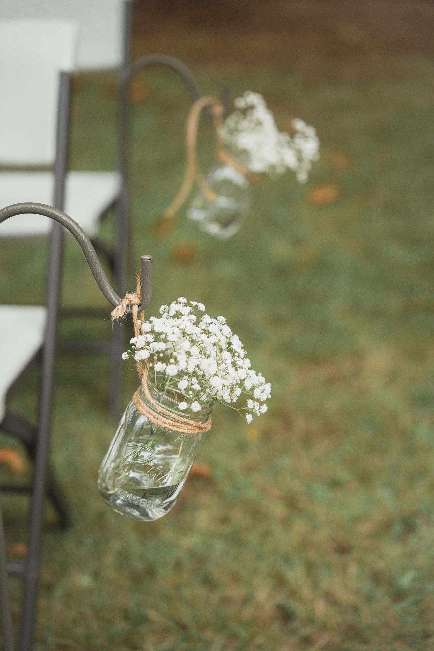 Mason jar with baby's breath flowers hanging on shepherd's hook at outdoor wedding ceremony aisle.