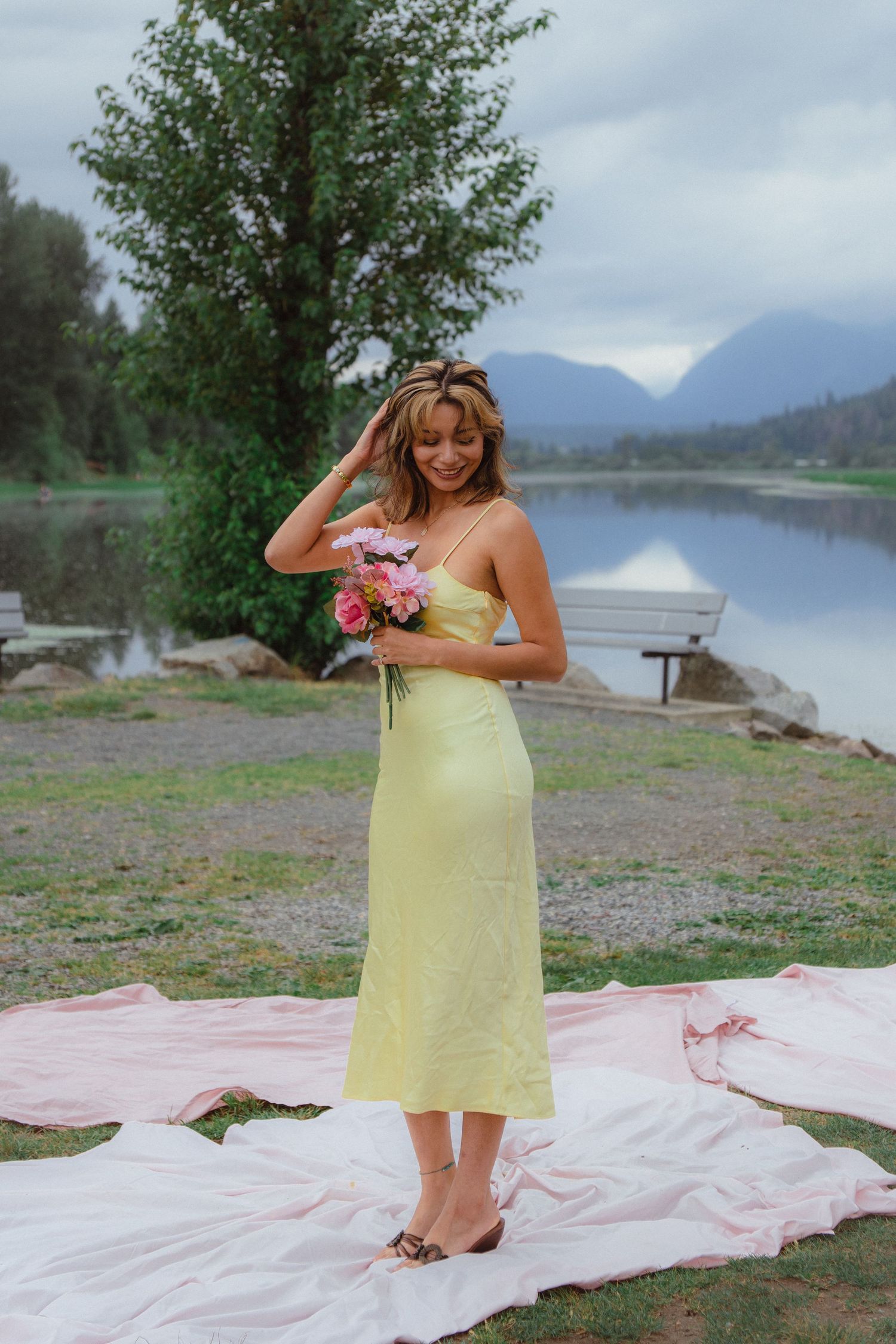 Woman in yellow satin midi dress adjusts hair while holding pink bouquet by mountain lake.