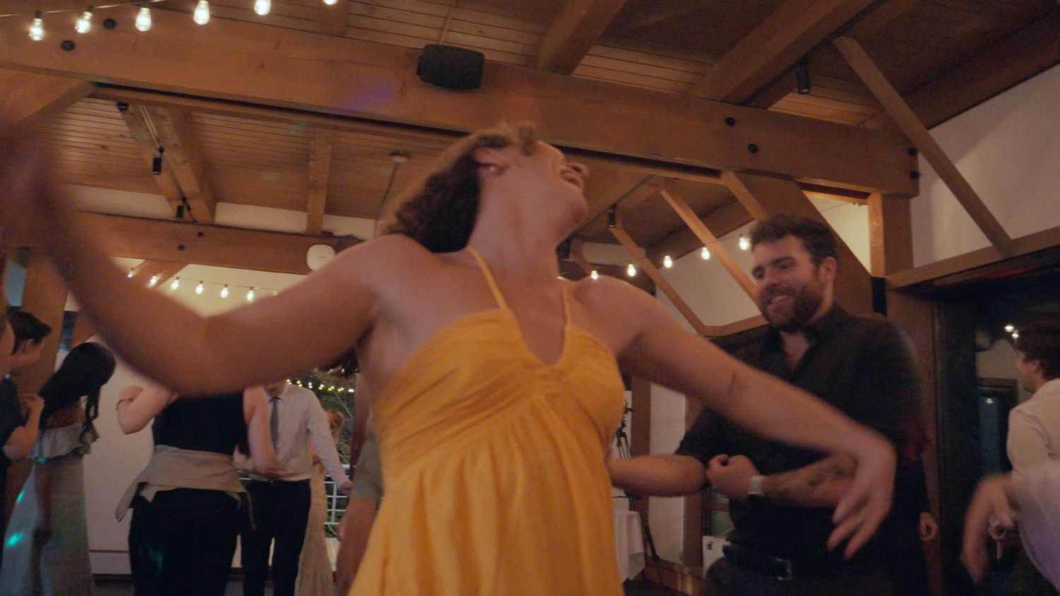 Woman in yellow dress dancing energetically at a wedding reception in a rustic venue with string lights.