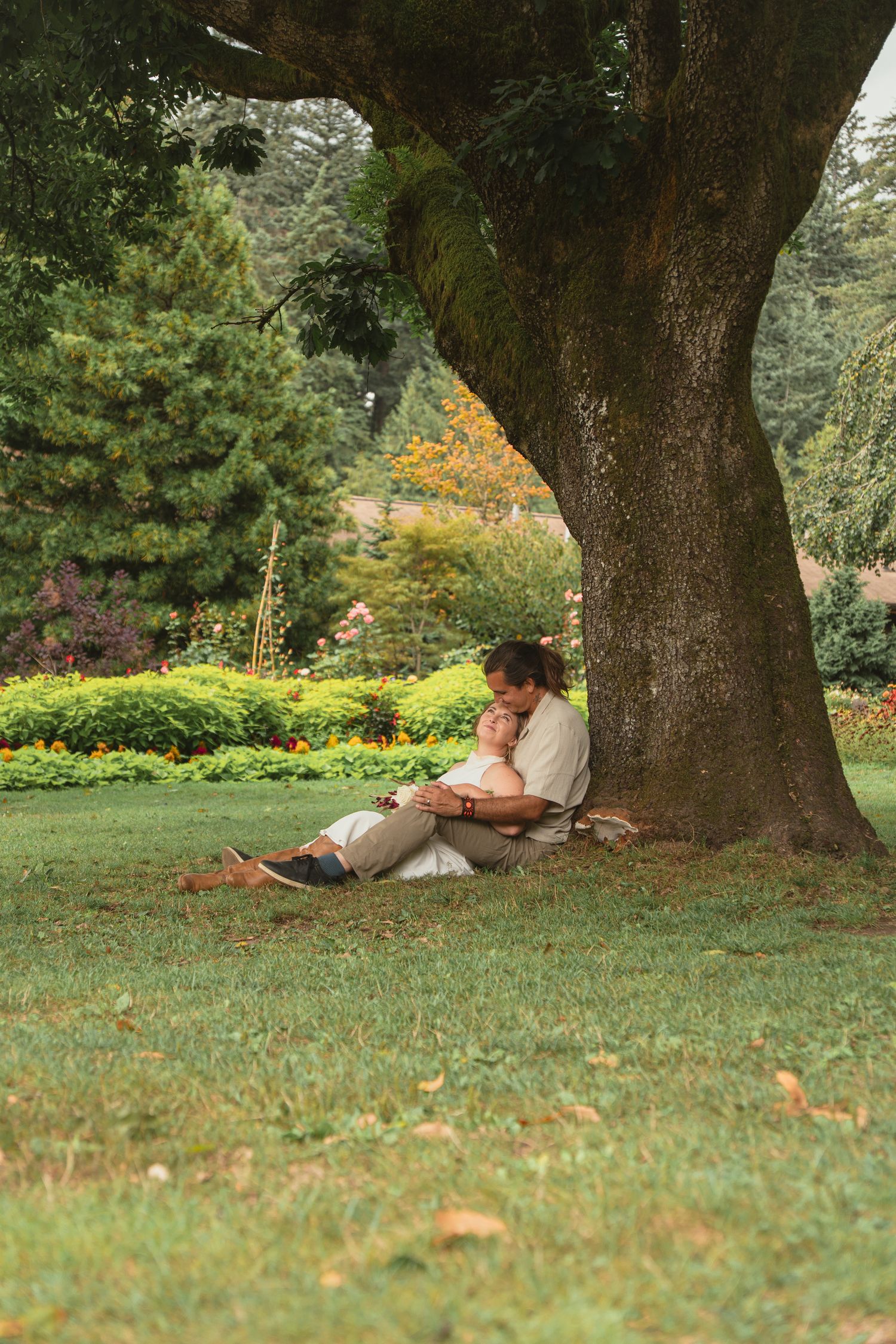 Newlyweds sit together under a large tree in a lush green park, sharing a quiet intimate moment.