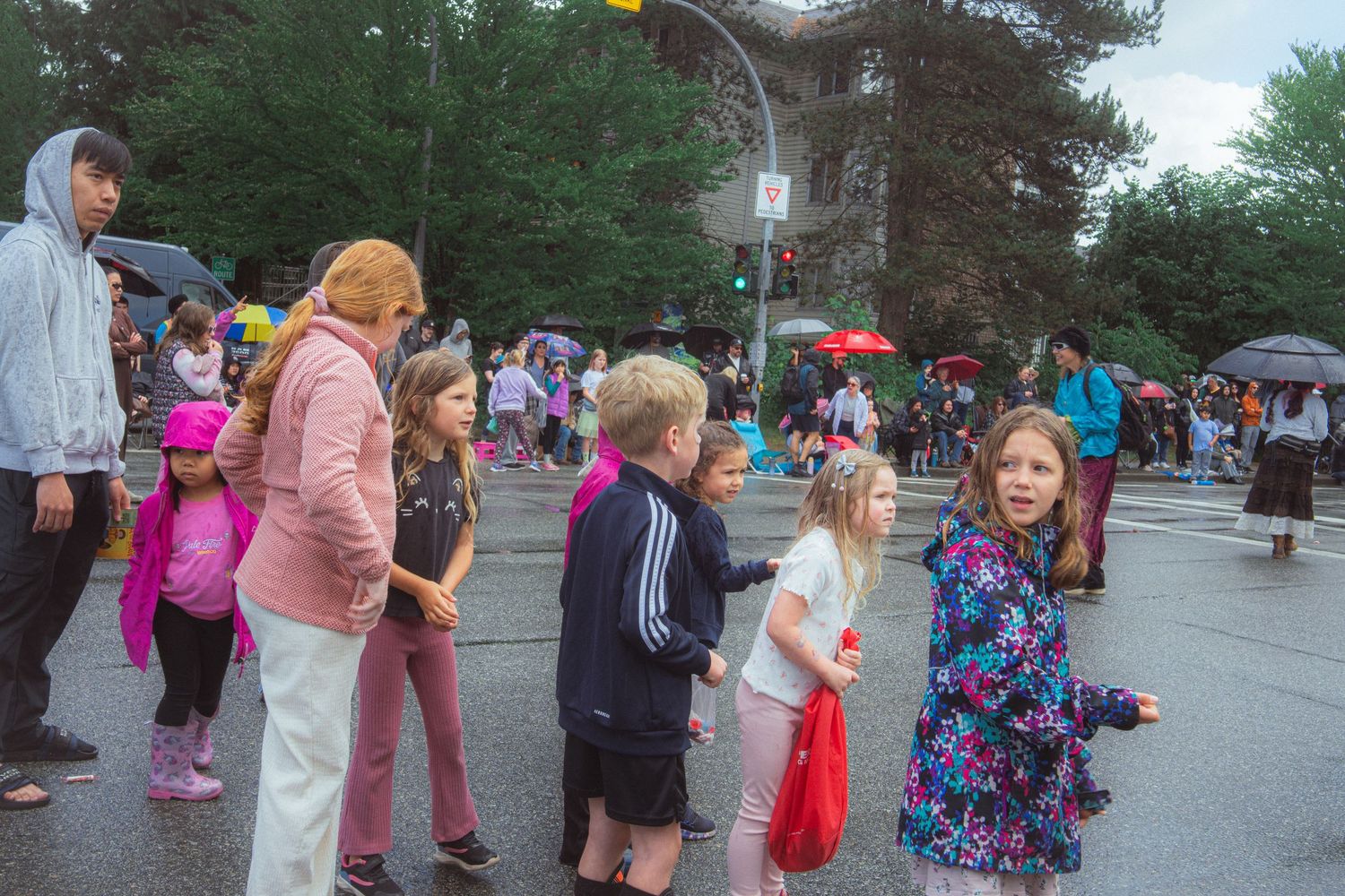 Children and adults standing on wet street during rainy outdoor community parade event.
