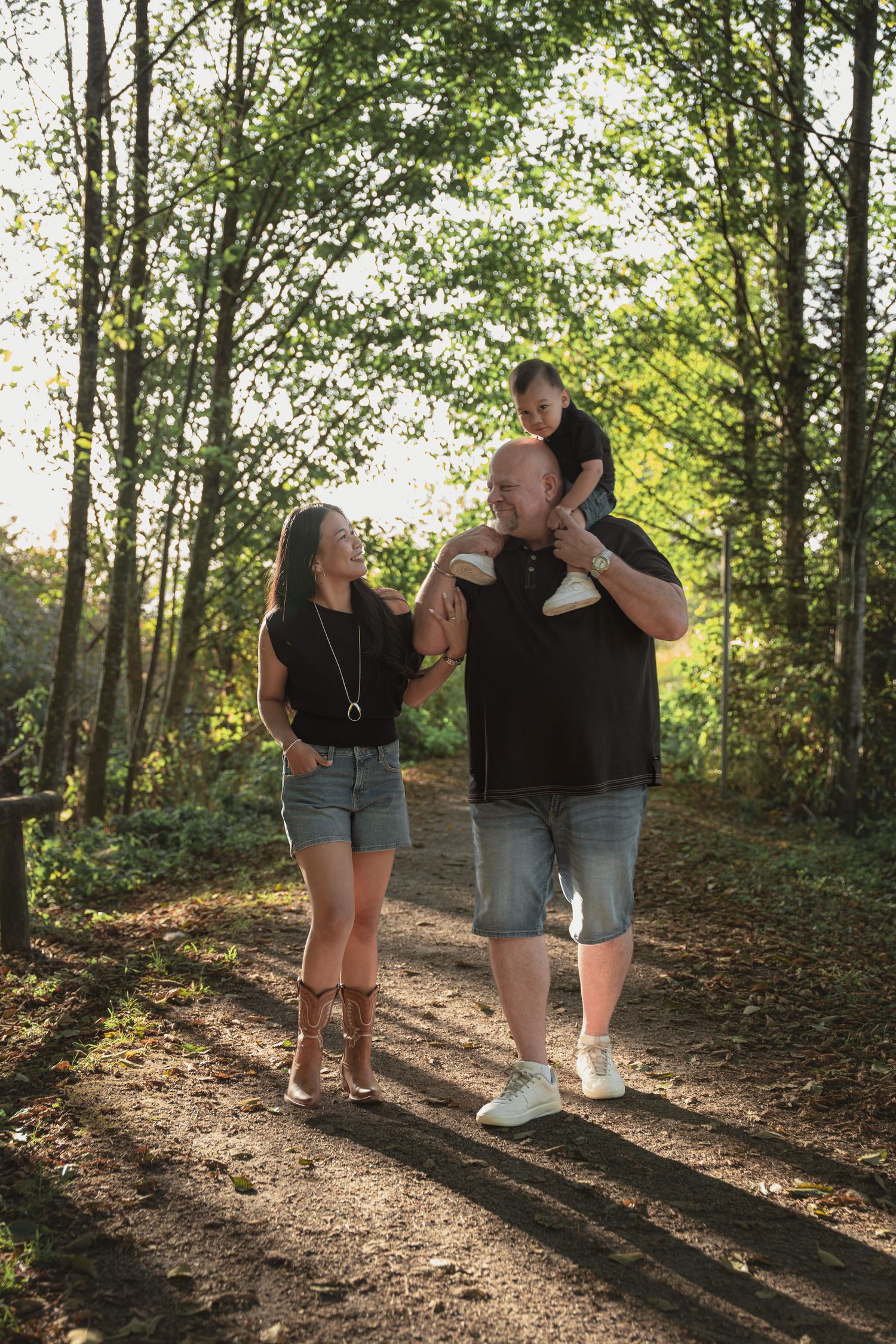 Family walking on a sunlit forest path, man carrying child on shoulders, woman smiling beside them.