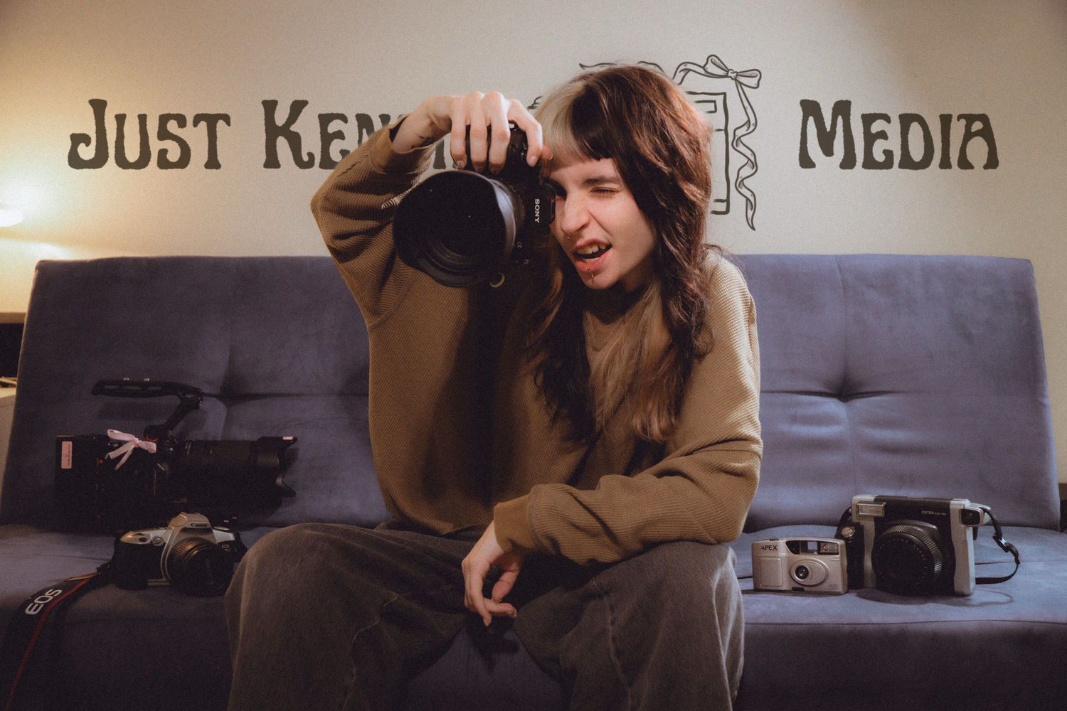 Woman holding a large camera lens on a couch surrounded by cameras, in front of a Just Kennedy Media sign.