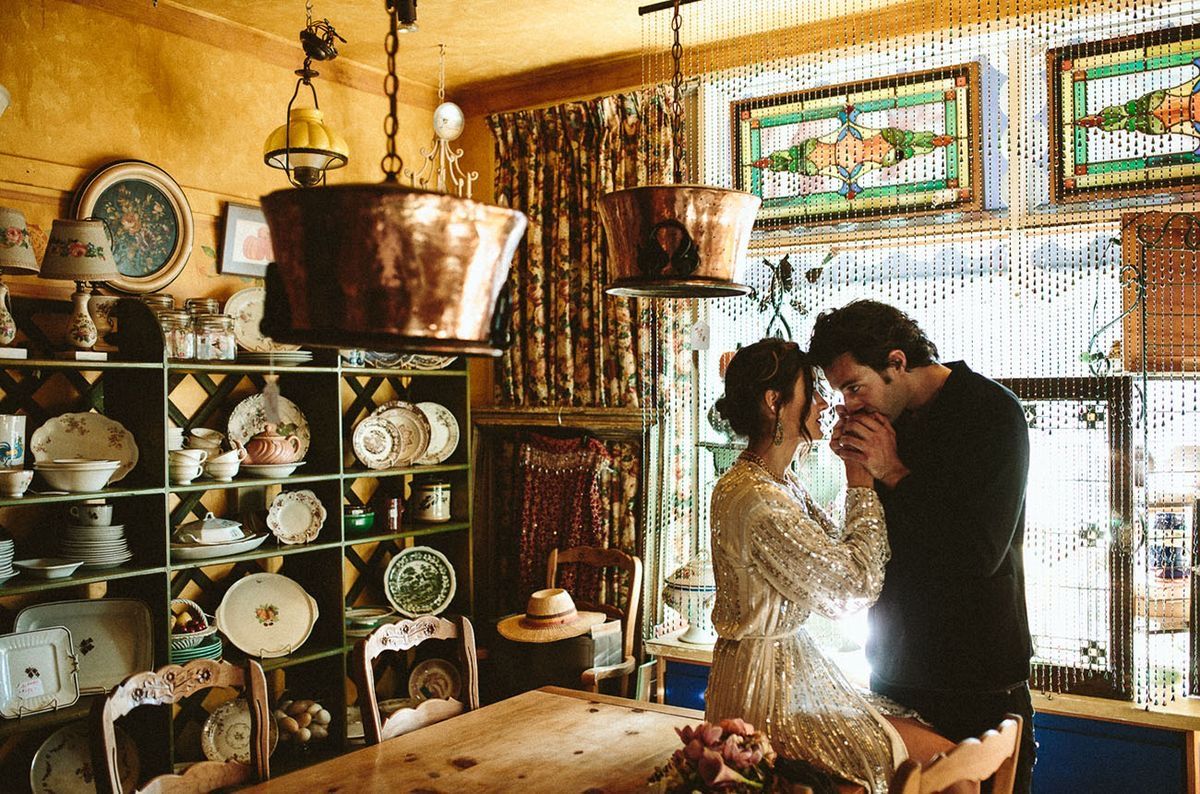 Couple sharing a romantic moment in a rustic antique shop filled with copper pots, ceramic plates, and stained glass windows.
