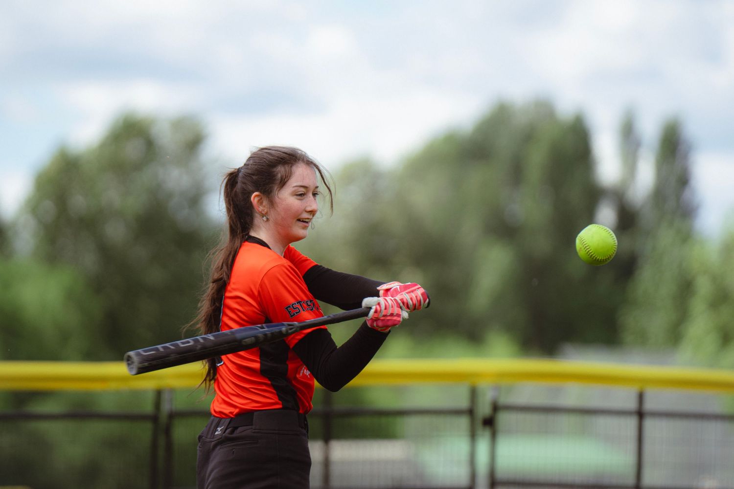 Female softball player in orange jersey making contact with softball outdoors.