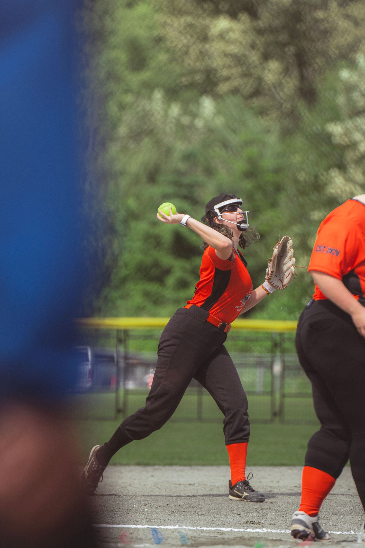Female softball player in orange uniform winds up to throw yellow ball during outdoor game.