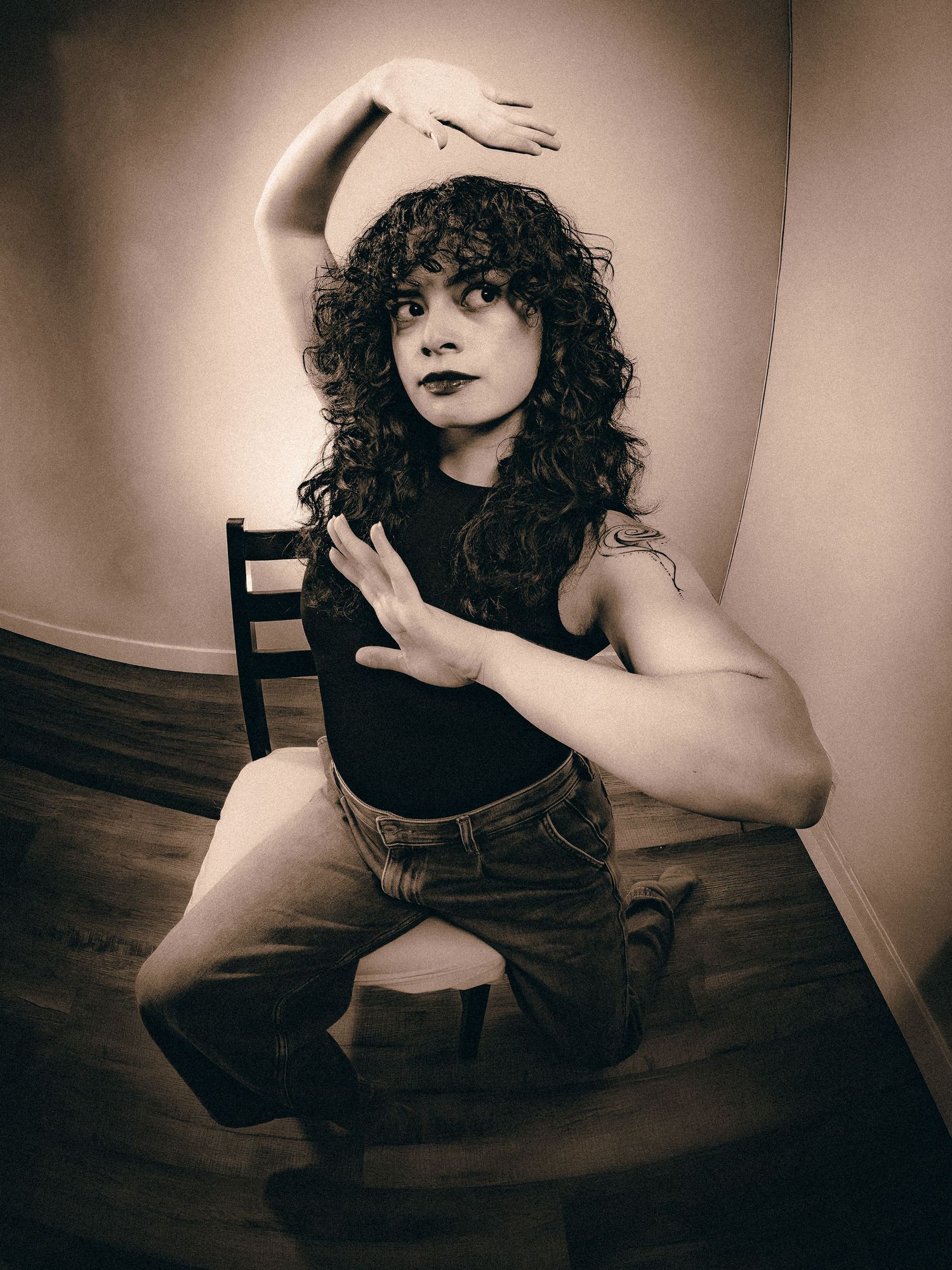 Woman with curly hair posing dramatically on a chair with expressive hand gestures in sepia tone.